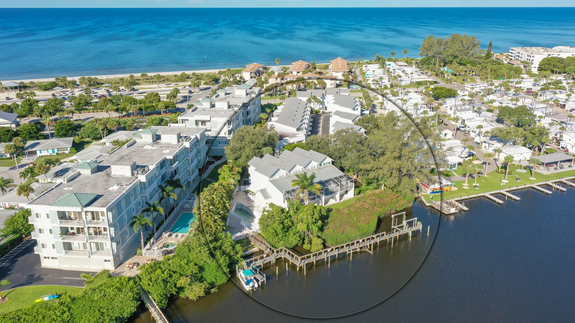 An aerial view showcasing a coastal community with residential buildings, palm trees, and a small marina near the shoreline. The vibrant blue ocean is visible in the background, highlighting the scenic coastal landscape.