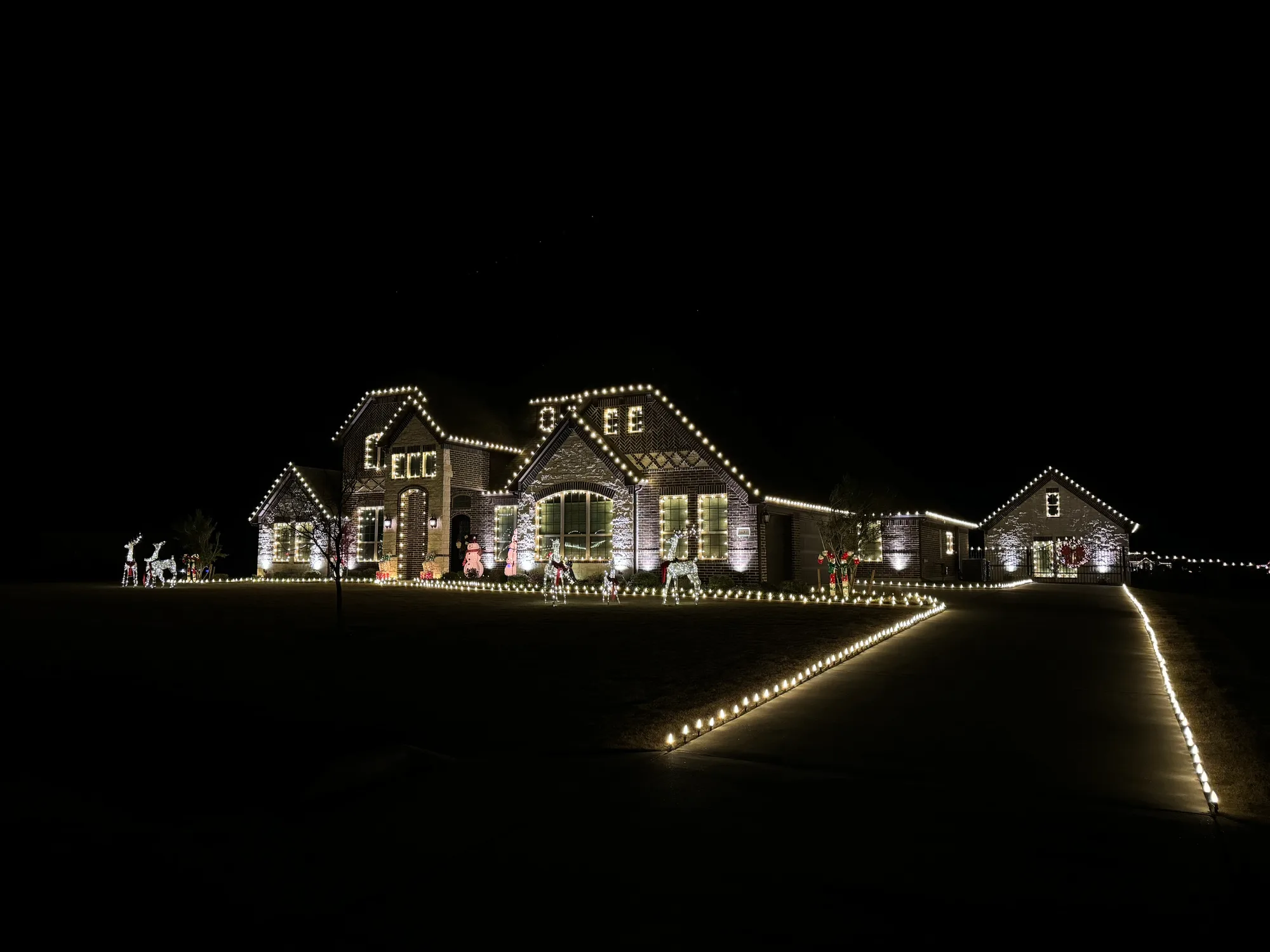 Large acreage property lit in warm white across the house, the separate outbuilding, the driveway stake lights and the garden beds, with lit reindeer on the lawn.
