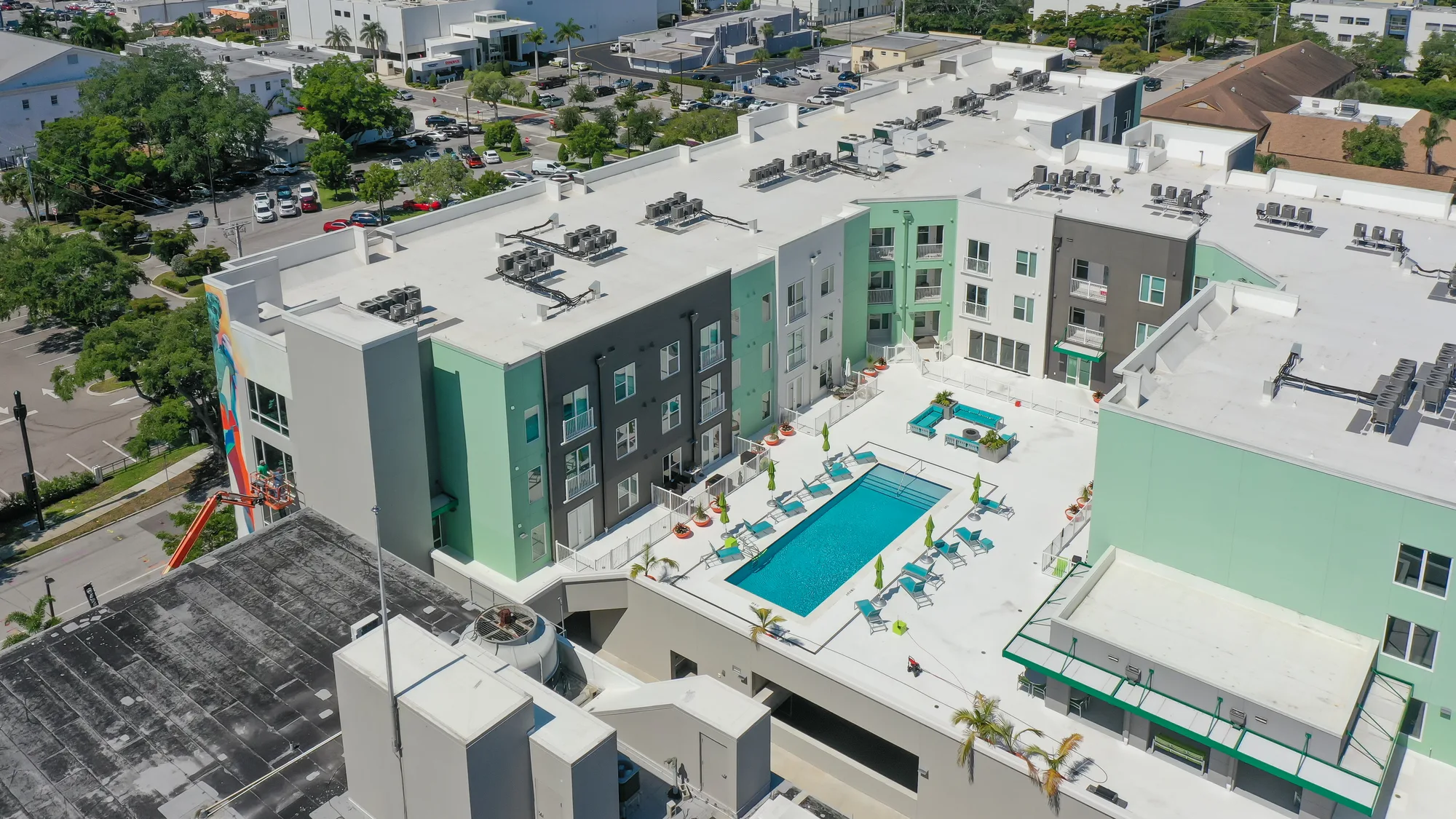 An aerial view of a modern apartment complex featuring a large swimming pool surrounded by lounge chairs. The buildings are in shades of green and gray, and construction work is visible on a wall nearby.