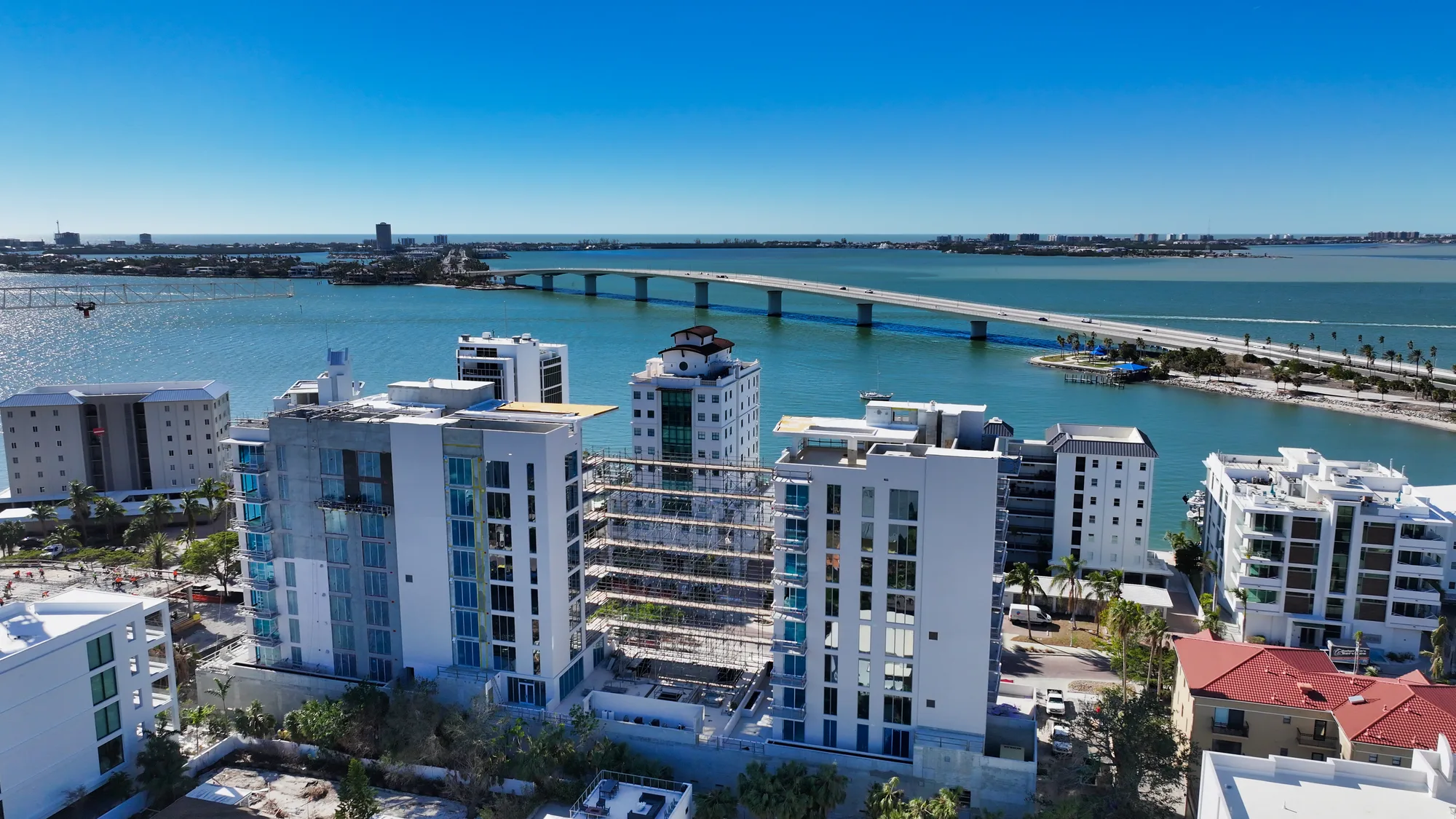 An aerial view showcasing modern coastal buildings alongside a turquoise body of water. A bridge spans the waterway in the background, with a clear blue sky overhead.