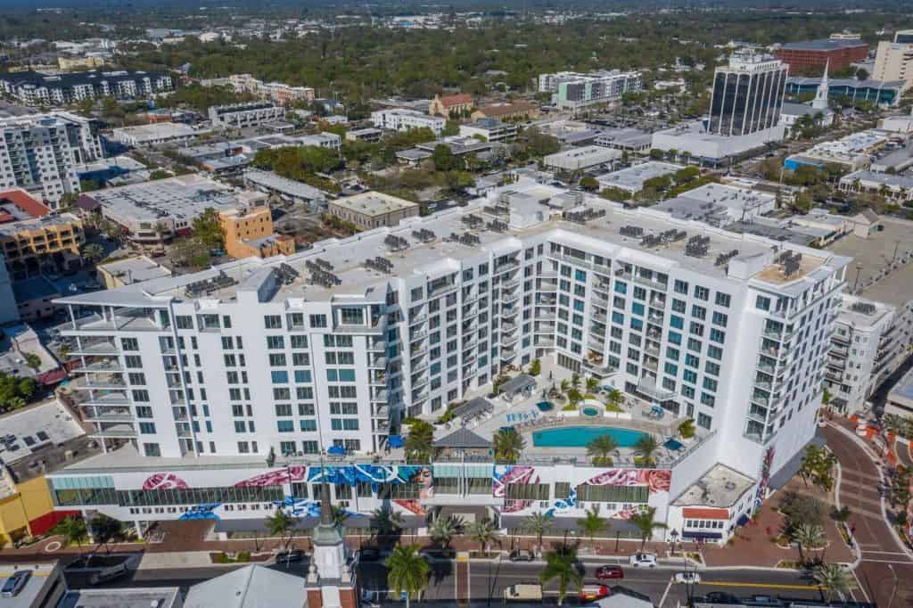 An aerial view of a contemporary hotel featuring a pool, located in a bustling urban area surrounded by various buildings and greenery. The hotel showcases a white facade with large windows and artistic decorations at its entrance.