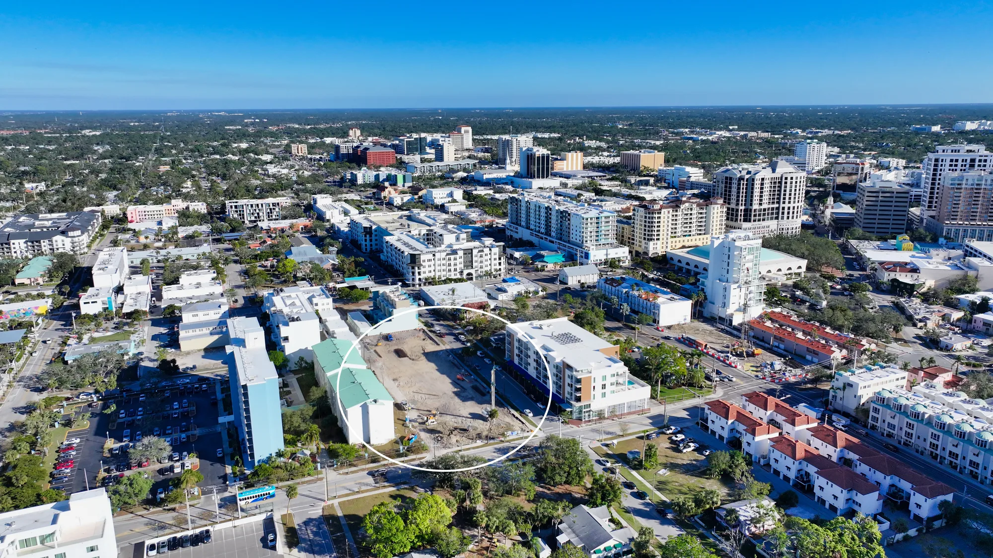This aerial image showcases a bustling urban area with a mix of residential and commercial buildings. An empty construction site is prominently featured in the foreground, indicating ongoing development amidst a backdrop of city structures and greenery.