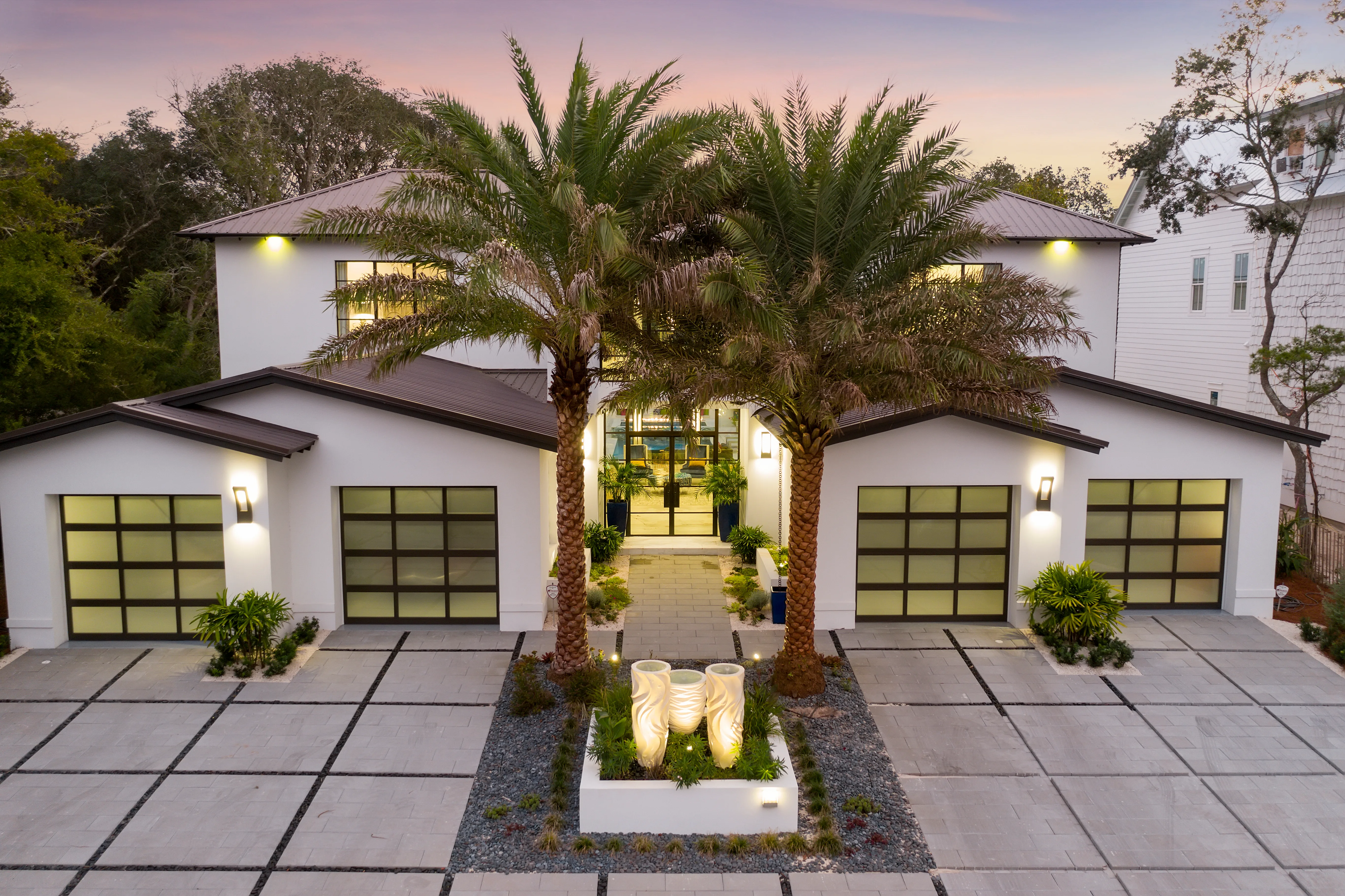 A finished modern home with tropical landscaping and lit garage doors.