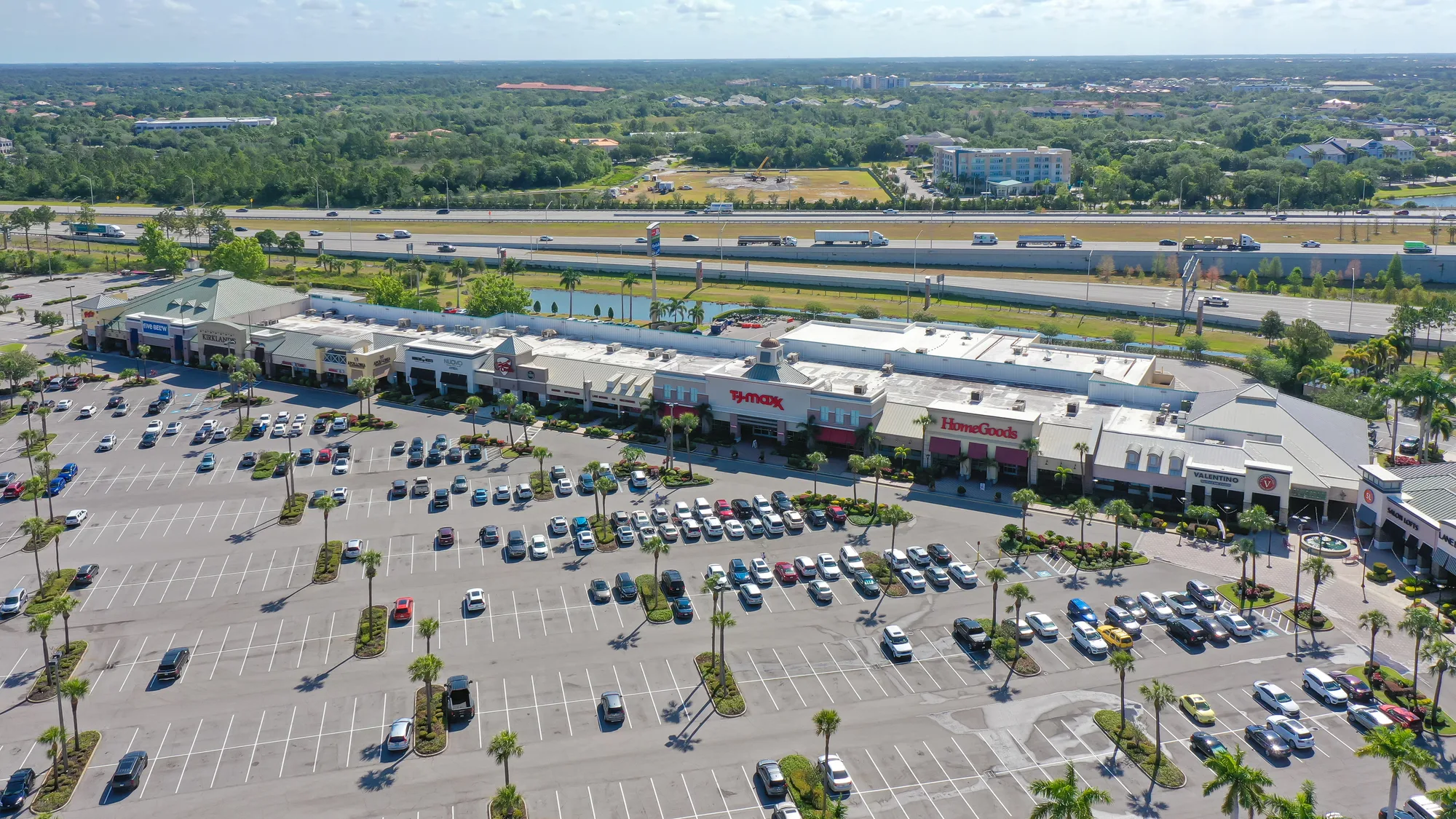 An aerial view of a shopping center featuring various retail stores, including HomeGoods and T.J. Maxx. The expansive parking lot is partially filled with cars, and a highway runs alongside the property.