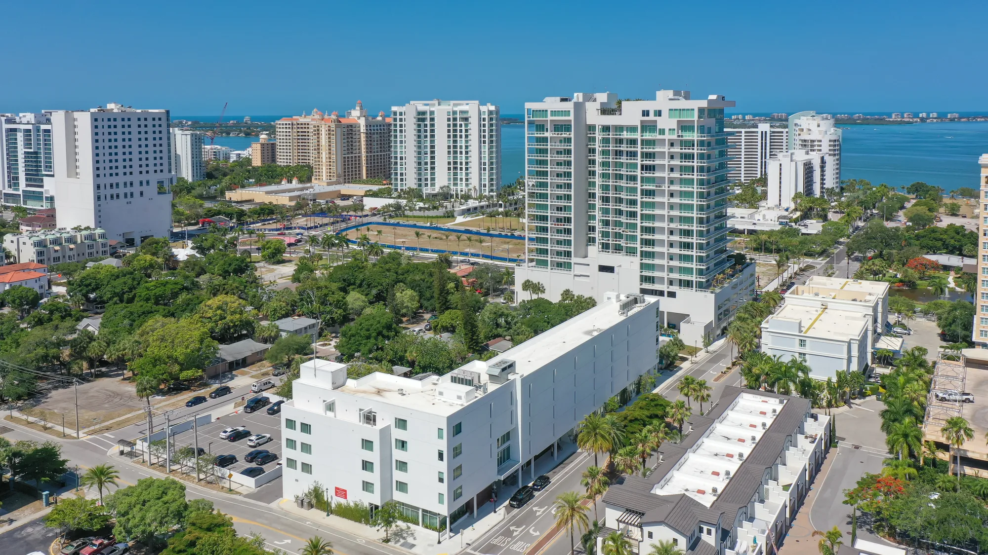This aerial photograph captures a coastal city featuring high-rise buildings alongside green spaces and palm trees. The image showcases a mix of urban development and natural landscapes with a clear blue sky and water visible in the background.