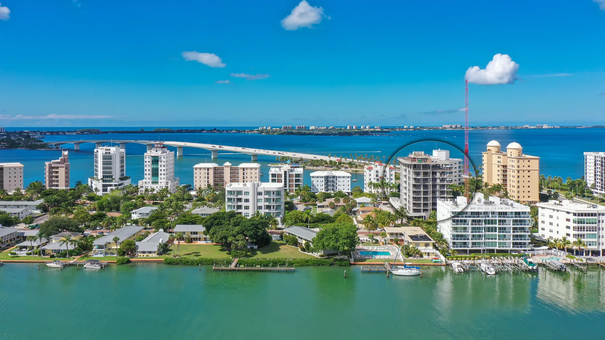 An aerial view of a coastal city featuring high-rise buildings, a marina with boats, and a bridge spanning across the water. The scene is set against a clear blue sky, showcasing greenery and residential areas along the waterfront.