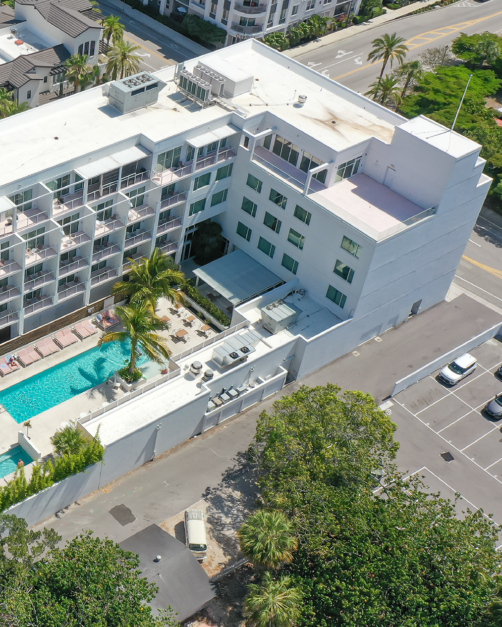 The image depicts an aerial view of a modern hotel featuring a swimming pool surrounded by lounge chairs and palm trees. Adjacent to the hotel are several parking spaces and nearby greenery, along with a roadway in the background.