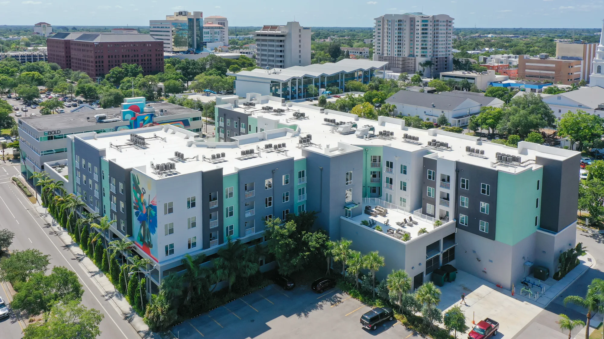 An aerial view of a modern apartment complex featuring colorful exterior design elements. The building is surrounded by greenery and urban developments, highlighting a blend of residential and commercial spaces in the city.