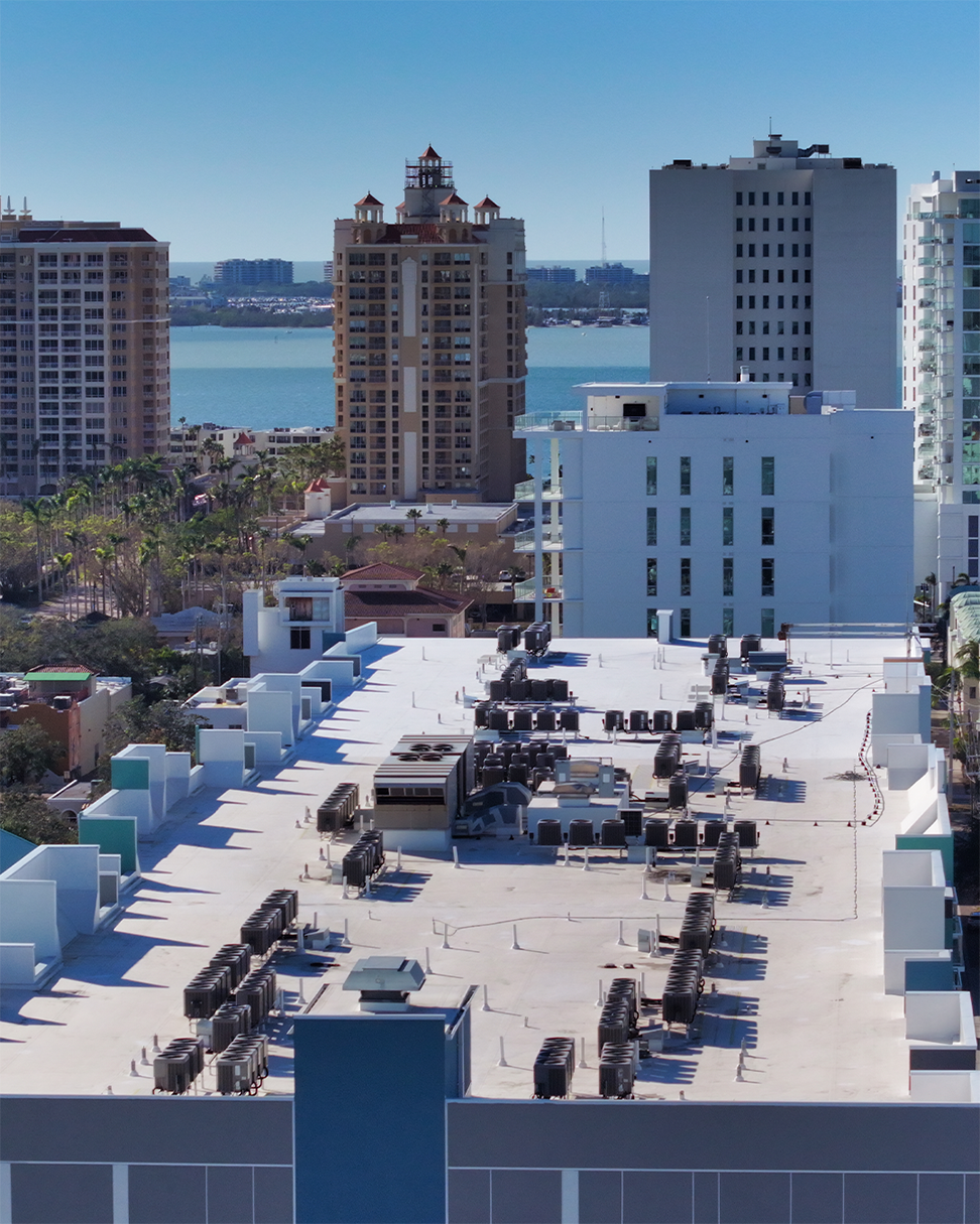 An aerial view of a rooftop with numerous HVAC units and machinery, surrounded by high-rise buildings. The backdrop features a view of a waterfront and distant city skyline under a clear blue sky.