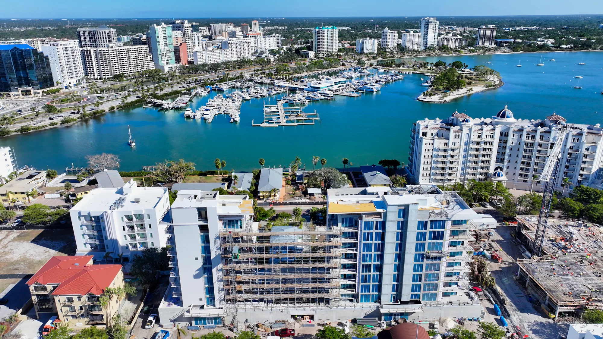 An aerial view showcasing a vibrant cityscape featuring a marina filled with yachts. The foreground includes several buildings, some under construction, surrounded by lush greenery and a clear blue waterway.