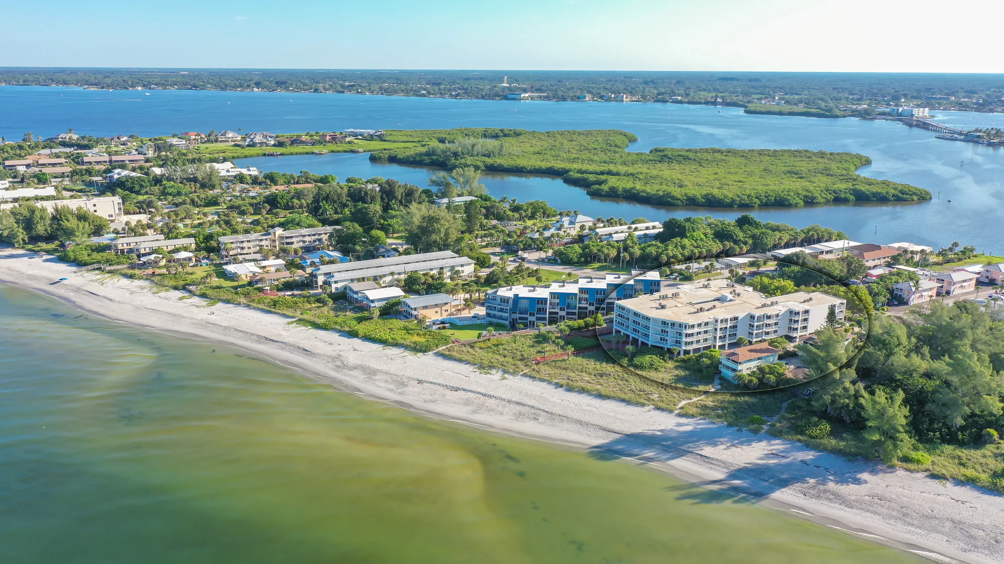The image shows an aerial view of a coastal area featuring residential buildings near a sandy beach, bordered by a calm blue waterway. Lush greenery and a mix of structures can be seen, highlighting the natural beauty and developed environment of the location.
