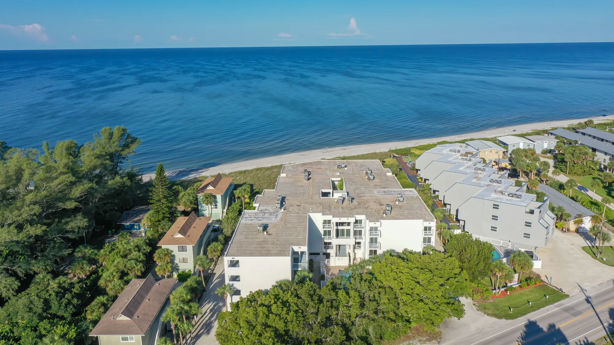 An aerial view of beachfront properties alongside a calm ocean. The image captures a mix of residential buildings and lush greenery, with sandy beach visible in the foreground.