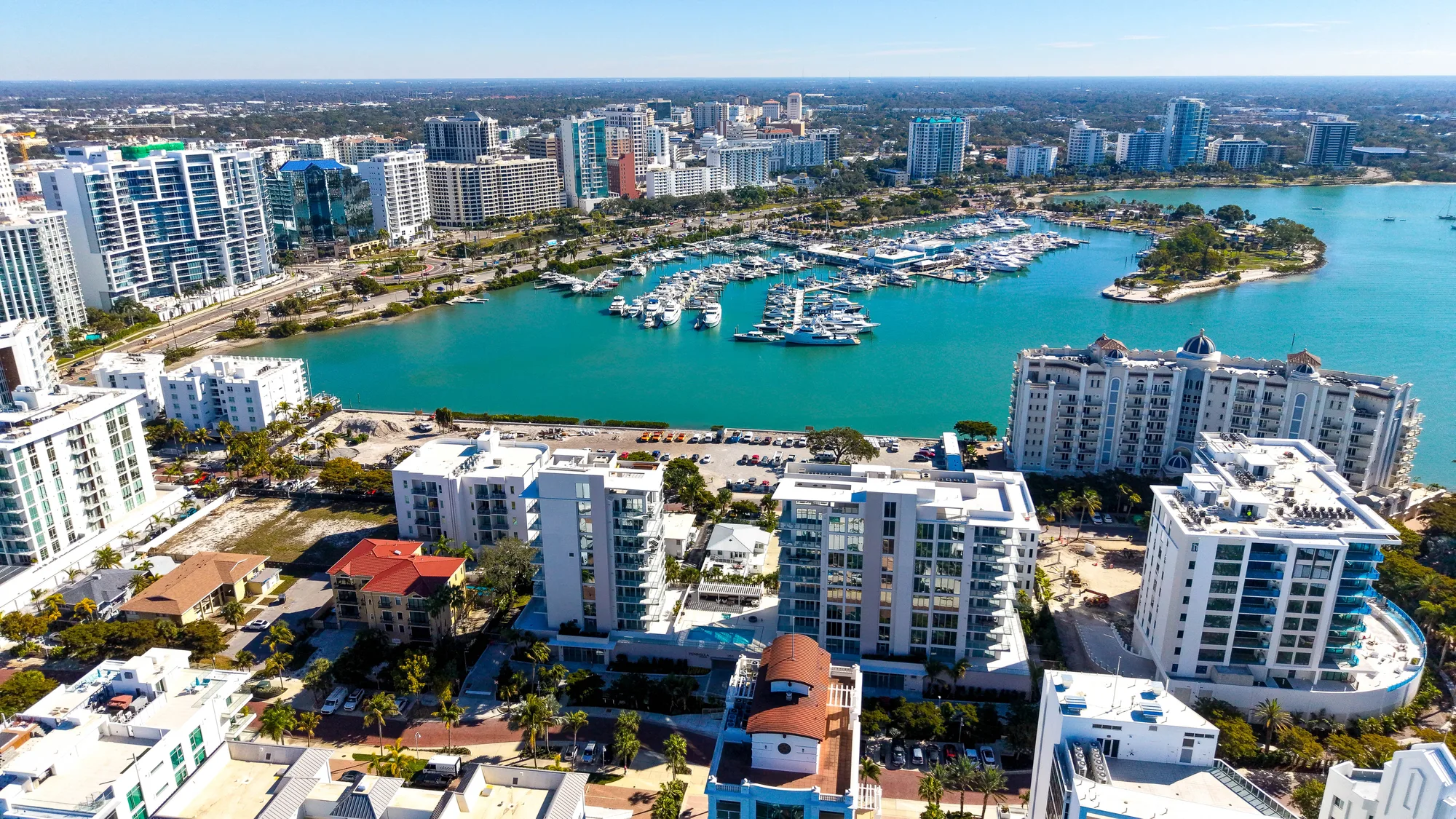 This aerial photo showcases a vibrant coastal city with modern high-rises overlooking a turquoise marina filled with yachts and boats. In the foreground, residential buildings and landscaped areas lead to the waterfront, highlighting the city's waterfront development.