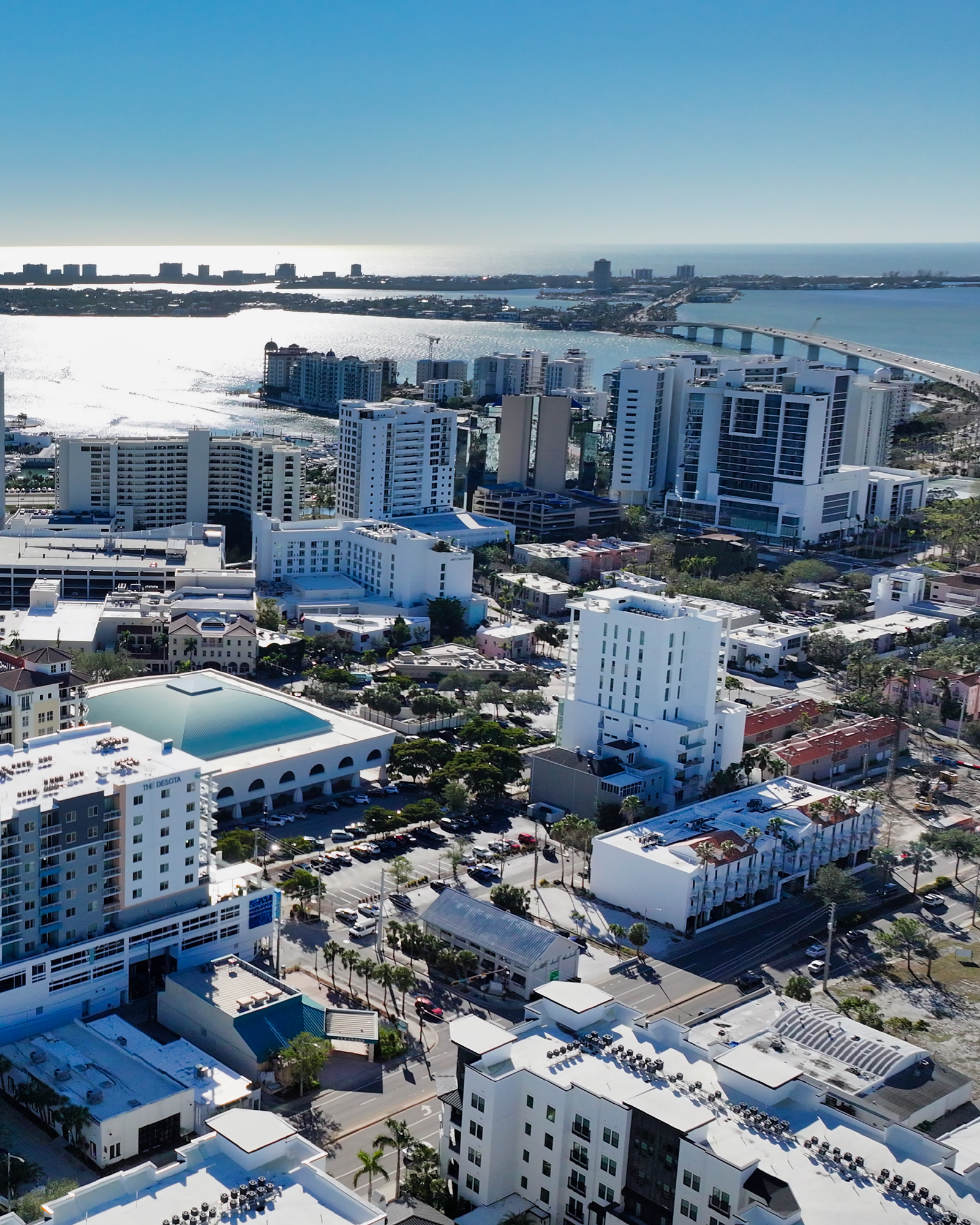 This aerial image showcases a vibrant coastal city with numerous high-rise buildings, streets, and waterfront views. The scene captures the blend of urban architecture and the shimmering ocean in the background, highlighting the city's dynamic landscape.