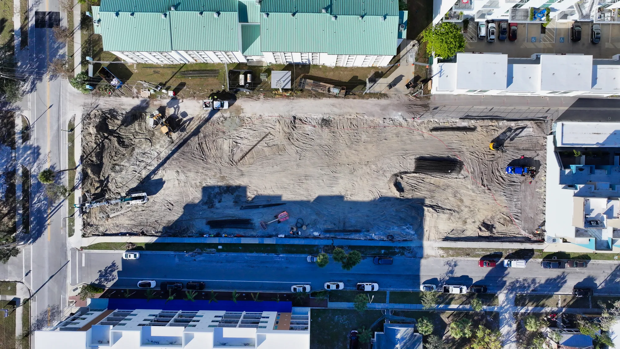 An aerial perspective shows an open construction site along with surrounding buildings and parked cars. Heavy machinery and workers are visible, indicating ongoing excavation and development activities.
