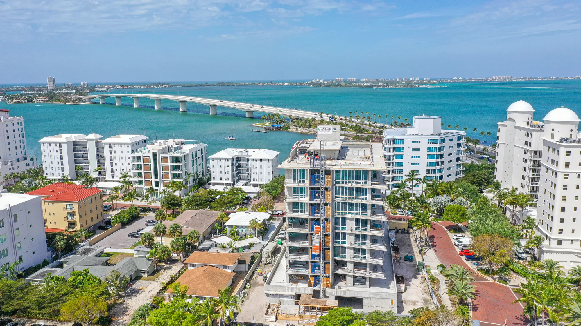 The image captures a coastal scene featuring a construction site of a multi-story building in the foreground, with residential buildings nearby. In the background, a bridge spans across turquoise waters, connecting the land, while palm trees and a clear sky enhance the tropical setting.