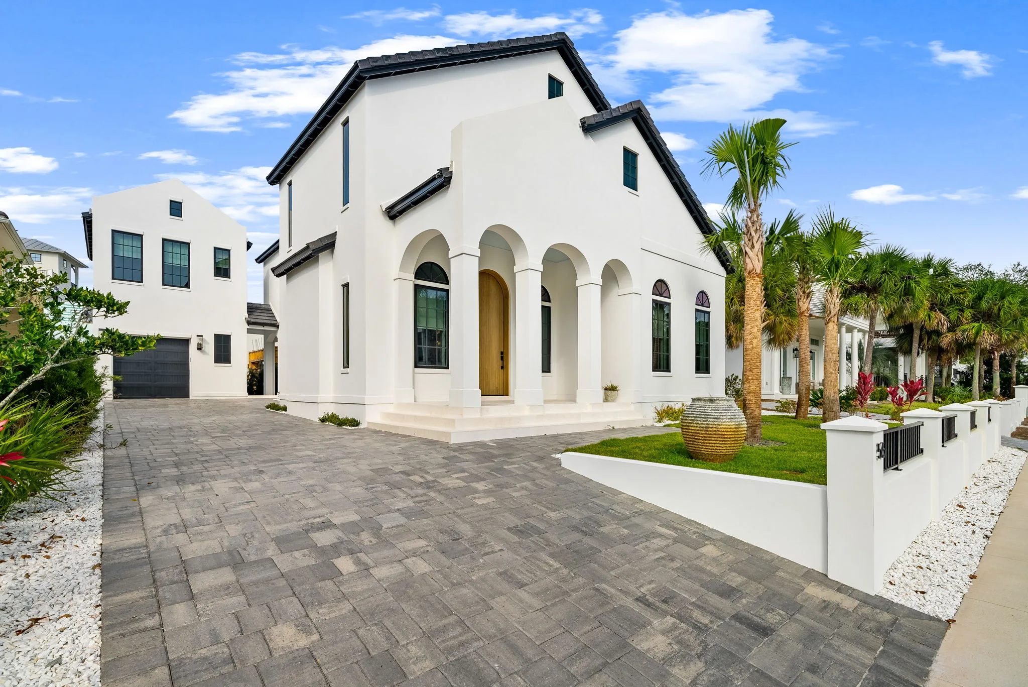 A white stucco house with arched entryways, palm trees and a paver driveway under a blue sky.