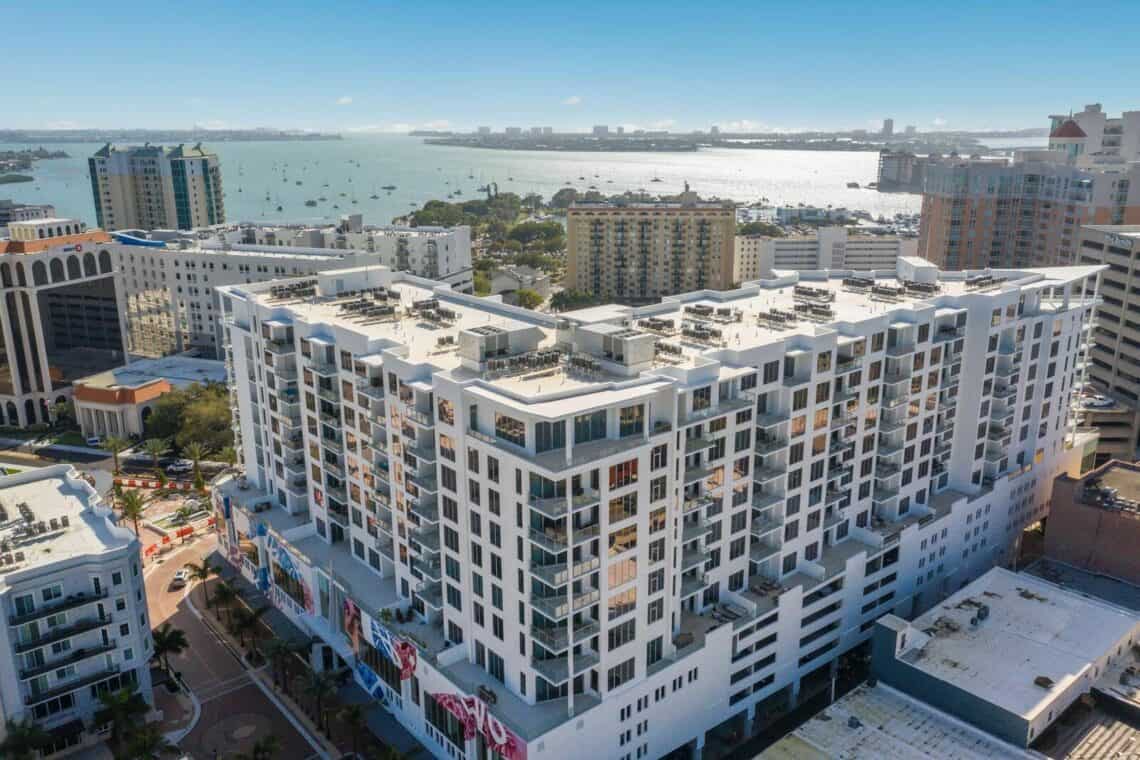 The image showcases a modern multi-story building in an urban setting with a waterfront view. The scene includes several nearby buildings and boats on the water, providing a glimpse of city life along the coast.