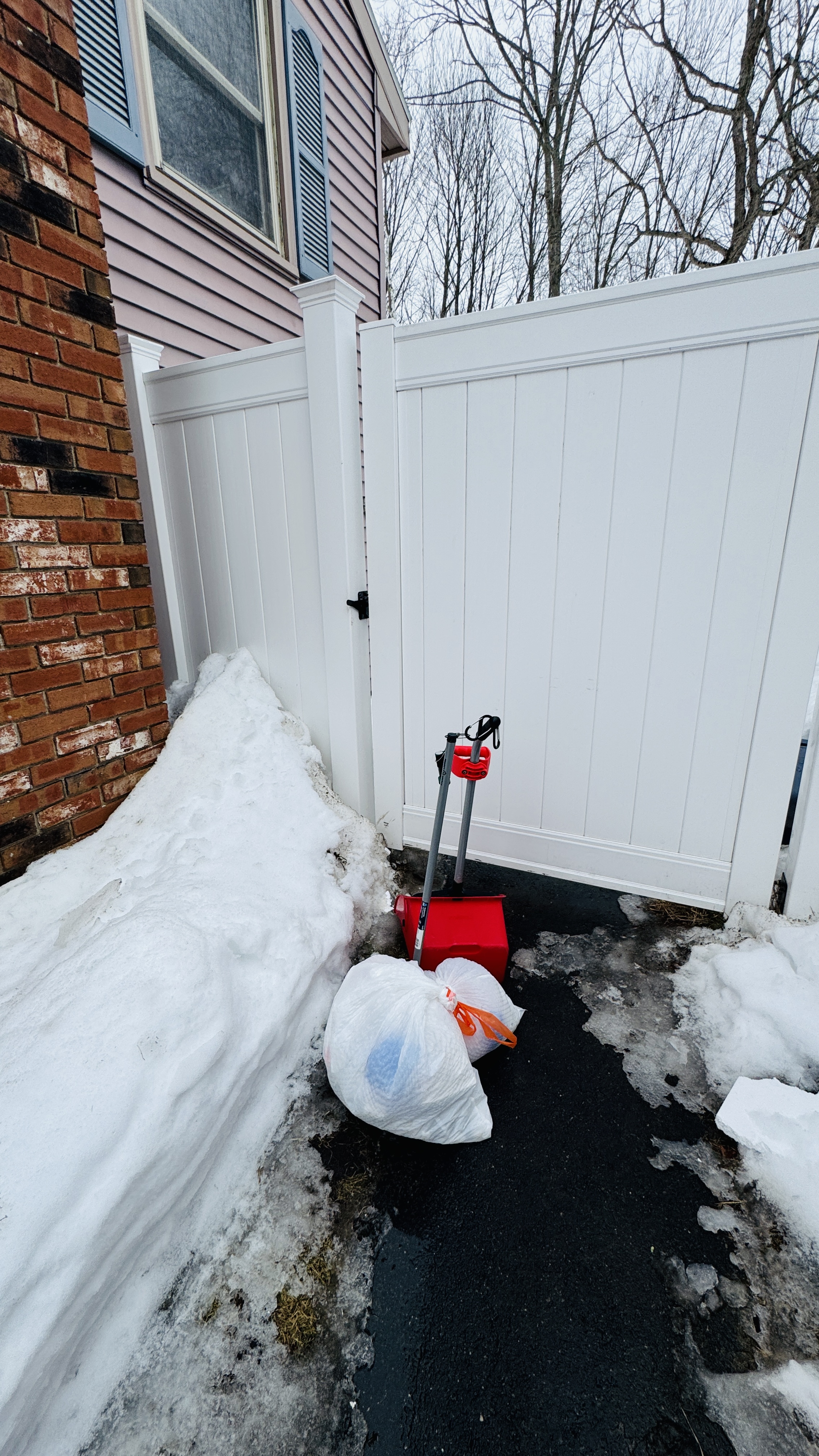Red shovel and a tied waste bag beside a house with snow still piled against a white fence
