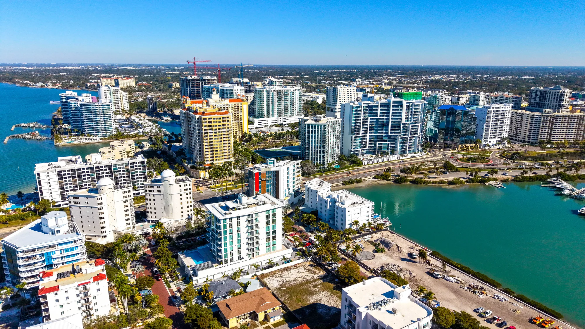 This aerial photograph captures a vibrant coastal city with numerous high-rise buildings alongside a blue waterfront. The image showcases the urban landscape, including buildings, palm trees, and a marina, under a clear blue sky.