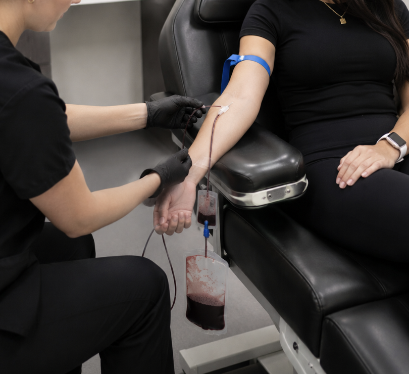 A blood donor seated in a medical chair with a healthcare professional preparing a blood draw.