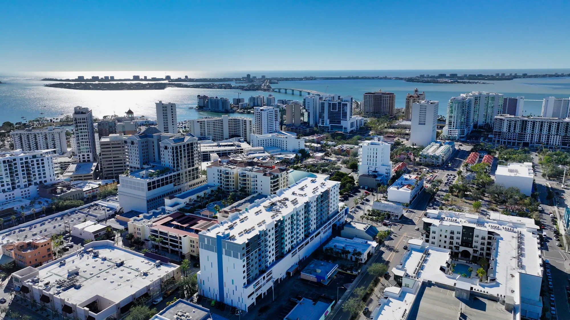 This aerial photograph showcases the skyline of Sarasota, Florida, featuring high-rise buildings along the waterfront. The image captures the shimmering water of the bay, with streets and residential areas visible in the foreground.