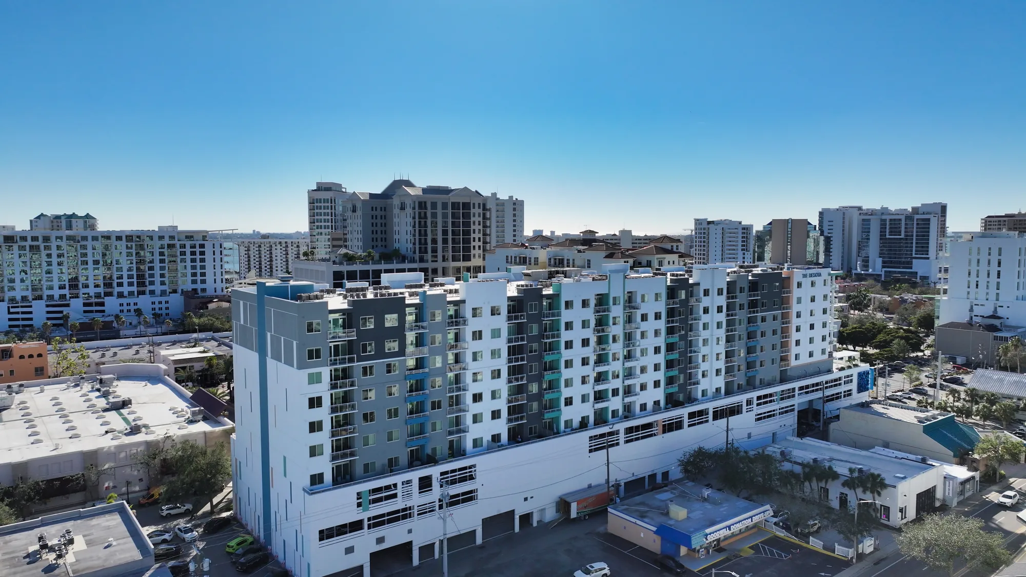 The image showcases a modern multi-story apartment building with a mix of white and gray facades, set against a backdrop of various urban structures. The scene captures a vibrant cityscape beneath a clear blue sky.