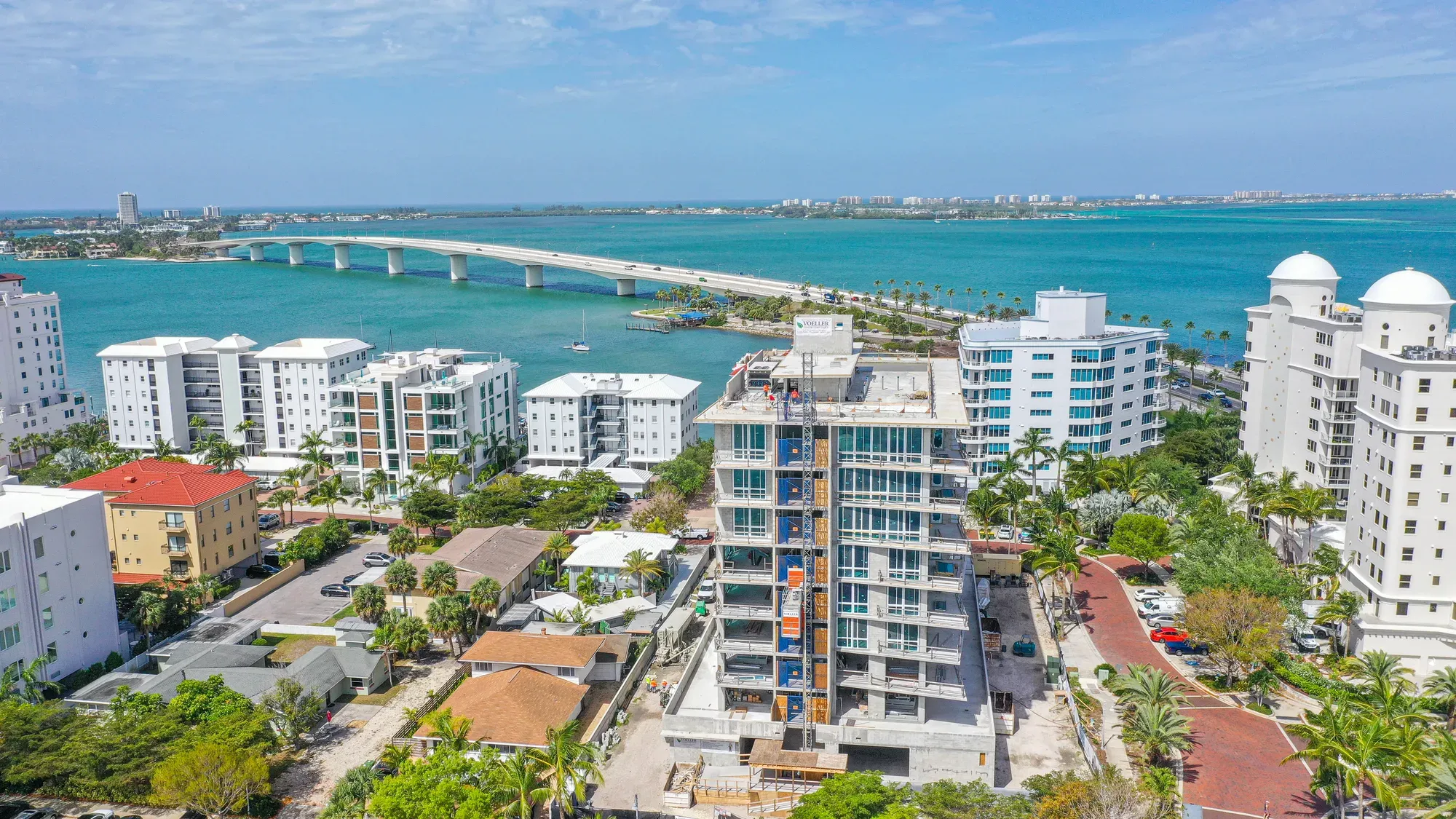 The image captures a coastal scene featuring a construction site of a multi-story building in the foreground, with residential buildings nearby. In the background, a bridge spans across turquoise waters, connecting the land, while palm trees and a clear sky enhance the tropical setting.
