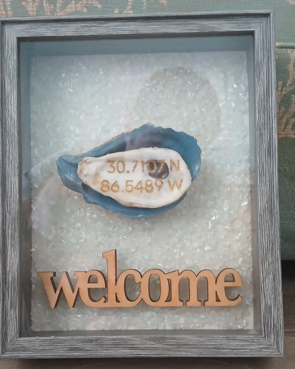 Gray shadow box holding an oyster shell engraved with map coordinates on a blue shell, above a wooden welcome sign