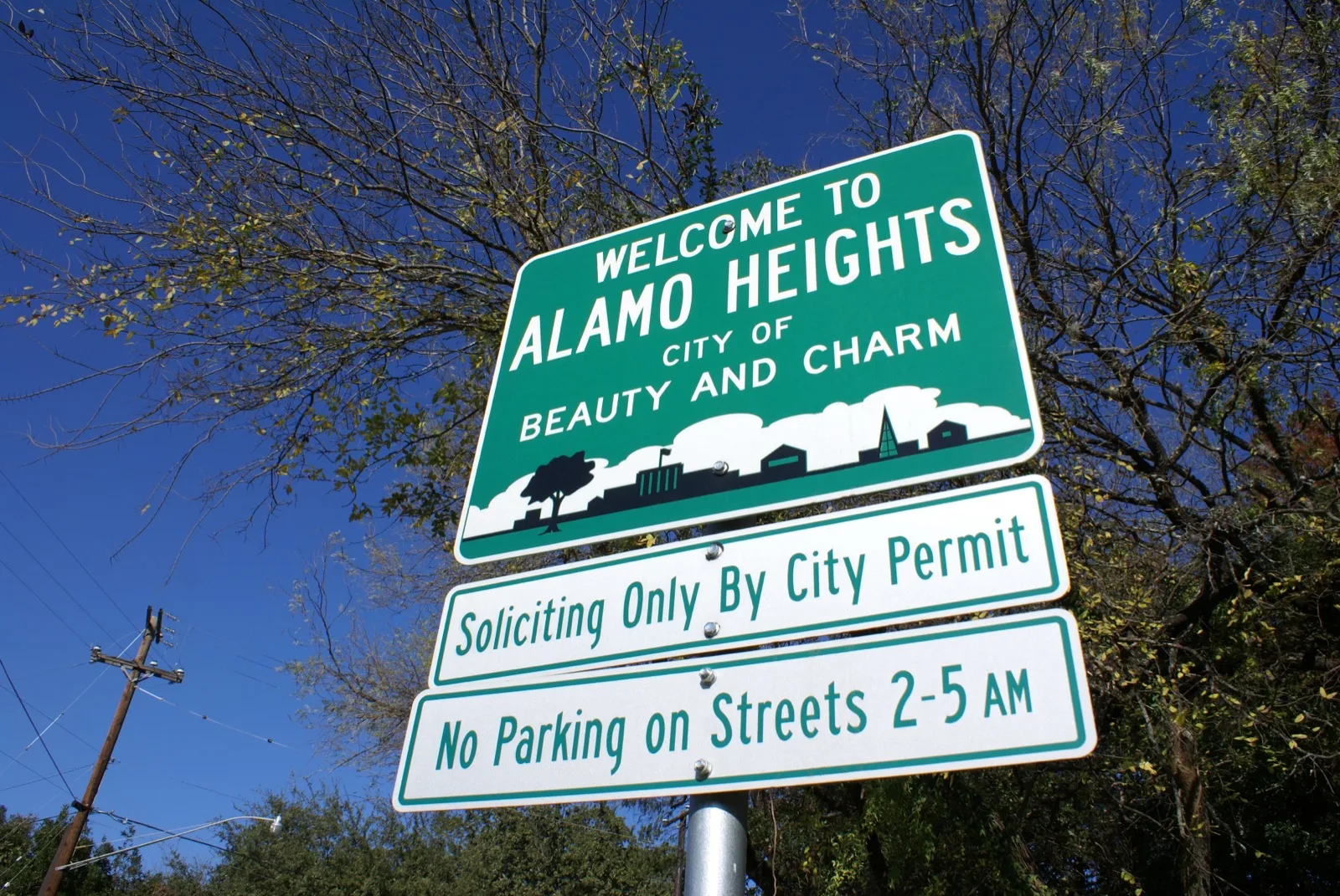 Green Welcome to Alamo Heights, City of Beauty and Charm sign under tree branches and a blue sky