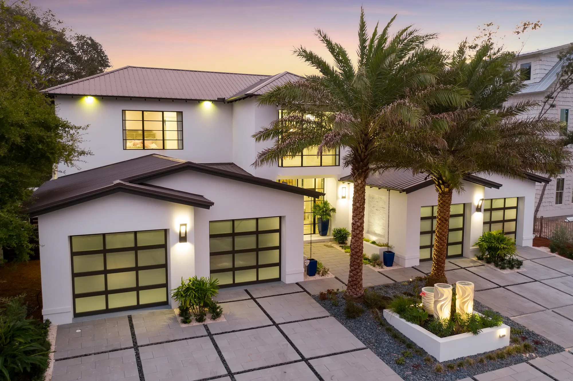 A finished home at dusk with lit glass garage doors and palm trees in the foreground.