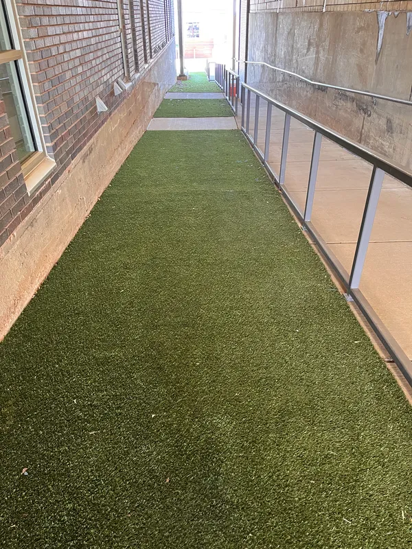 Narrow artificial turf pathway between brick walls with a railing on one side leading towards an open area