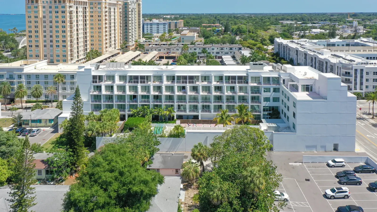 Aerial view of a modern coastal apartment complex featuring multiple floors and balconies. The image showcases surrounding greenery, parking areas, and nearby taller buildings along a coastal landscape.