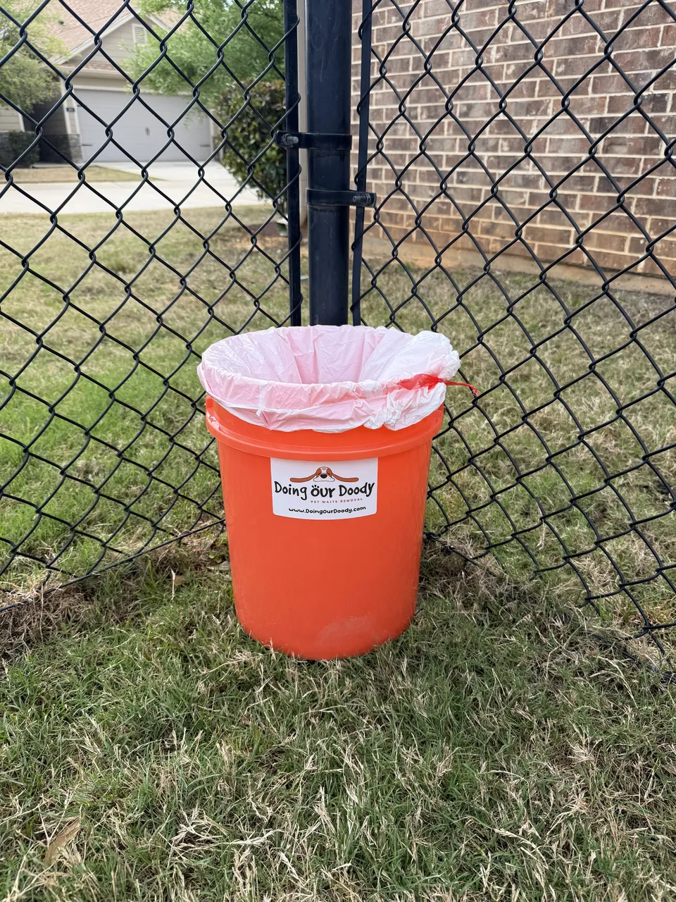 Orange Doing Our Doody pet waste disposal bin next to a black chain-link fence
