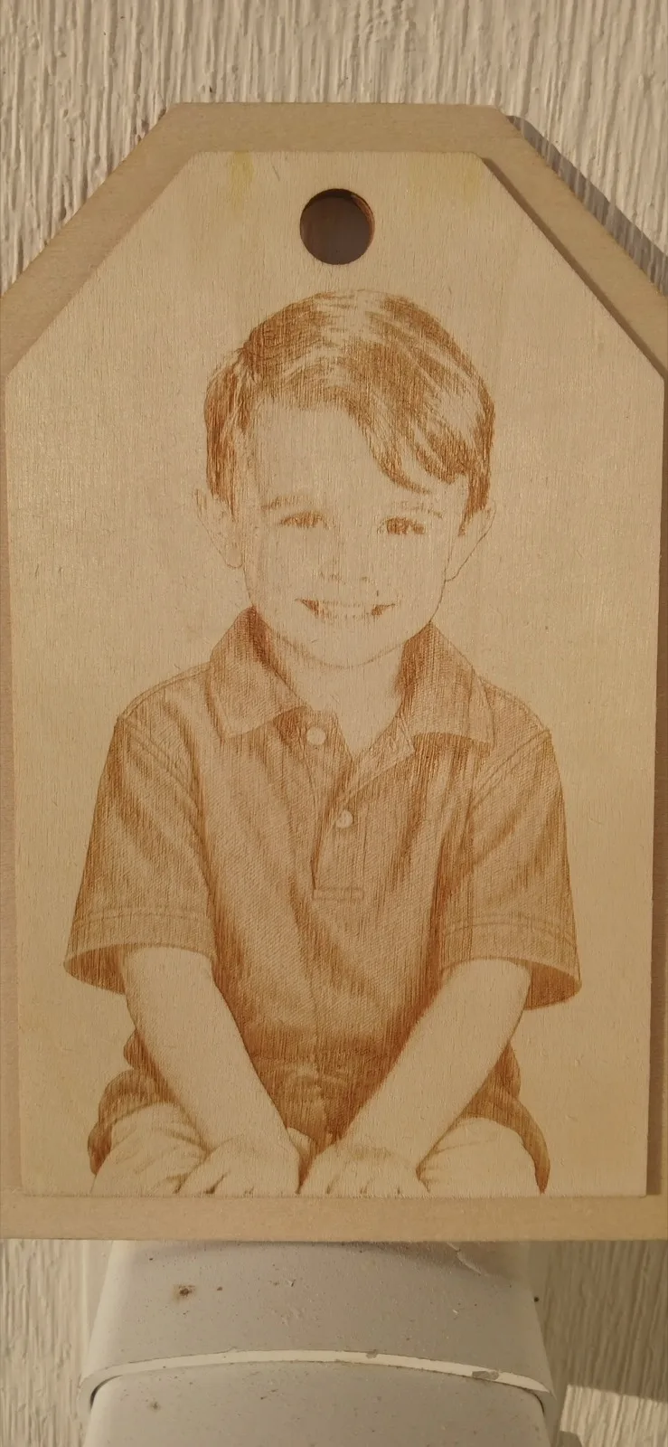 Wooden gift tag engraved with a portrait of a smiling young boy, displayed against a light wall
