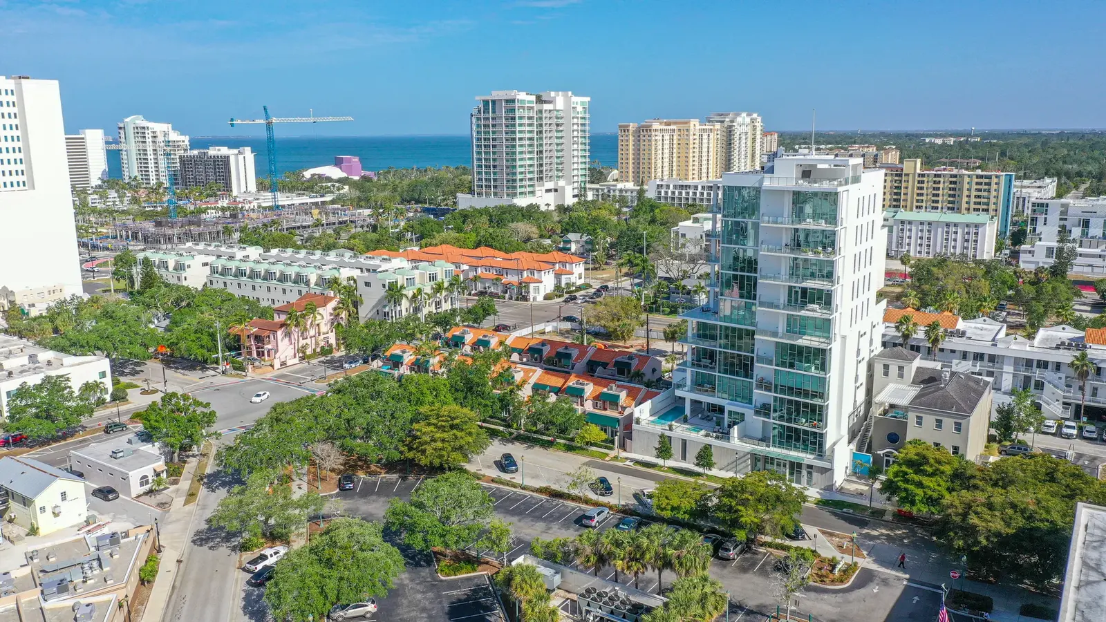 Aerial of The Collection Residences in Sarasota, a white multi-storey residential building among the downtown low-rise blocks, with the Gulf beyond.