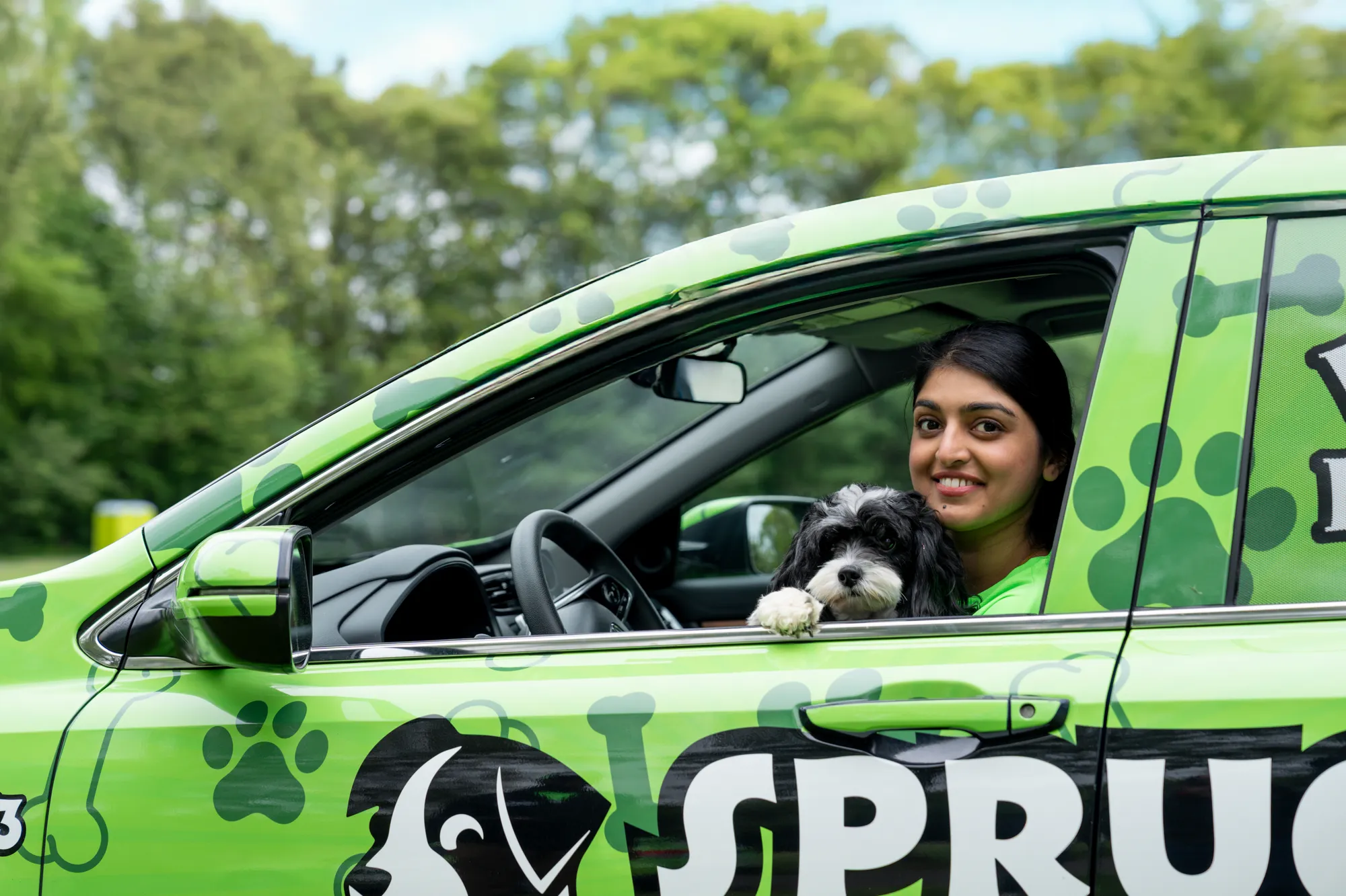 A smiling Spruce Scoop team member in the driver's seat of a wrapped Spruce Scoop car with a small black-and-white dog at the window