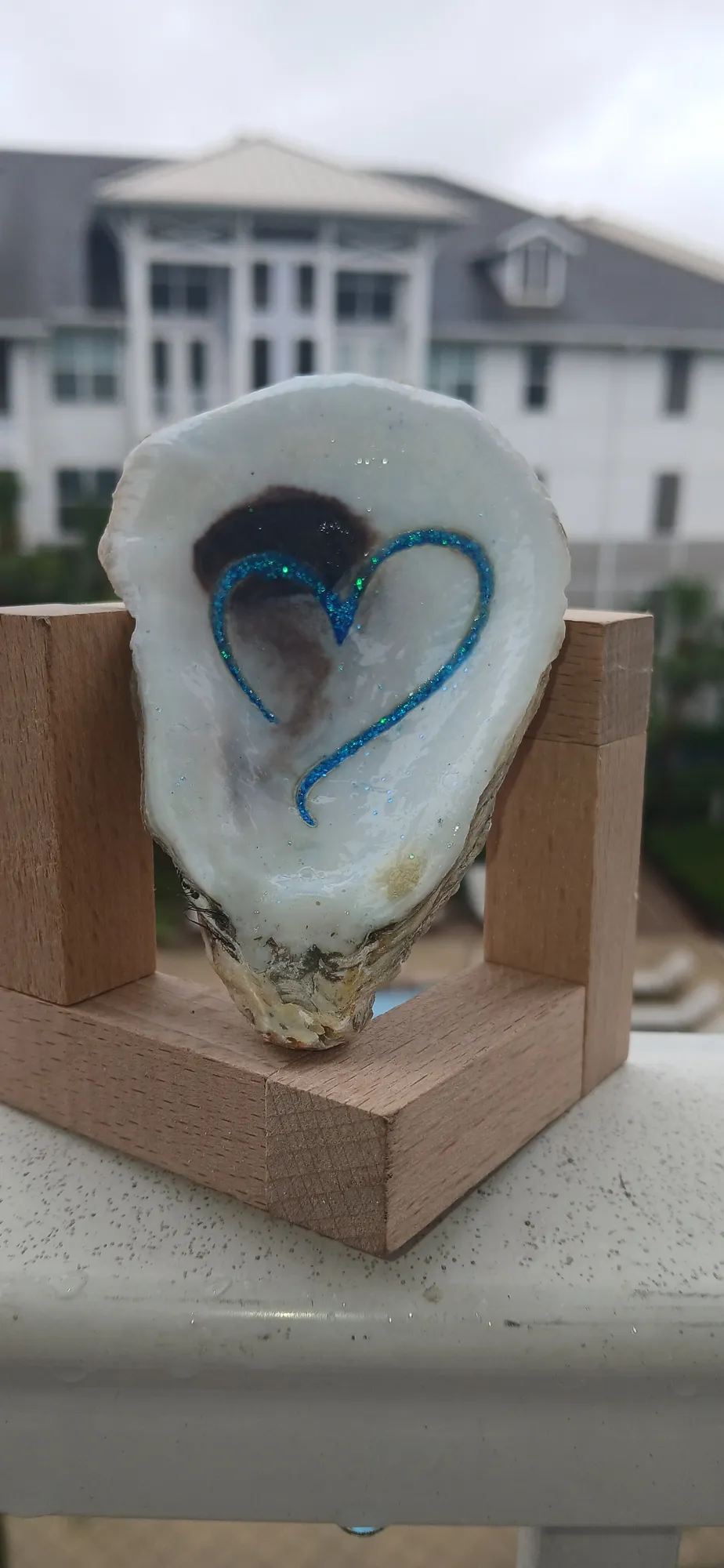White oyster shell with a blue glitter heart, standing in a wooden holder on a balcony rail