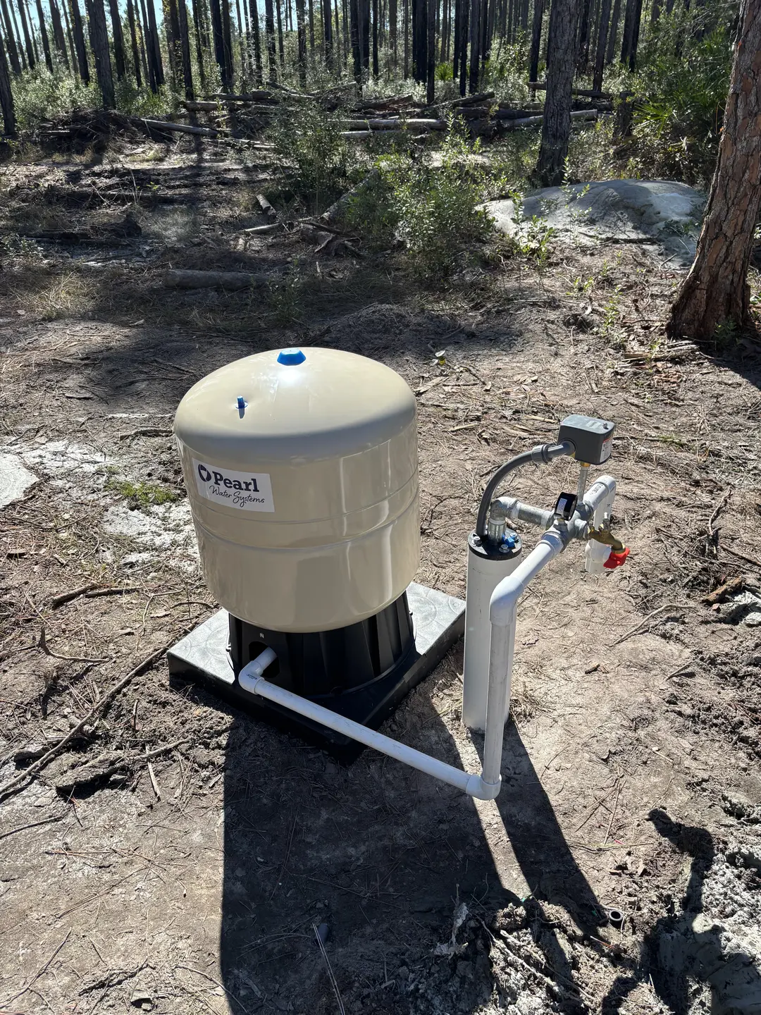 Beige Pearl Water Systems pressure tank with connected piping mounted on a black base in a forested area