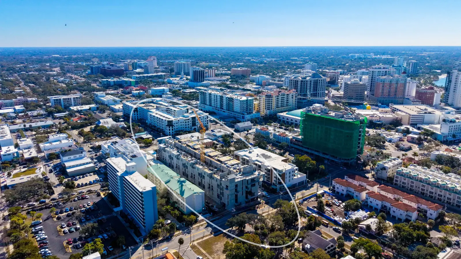 This aerial image captures a bustling urban landscape featuring a mix of commercial and residential buildings, with new construction evident in the foreground. The backdrop shows a city skyline under clear blue skies.