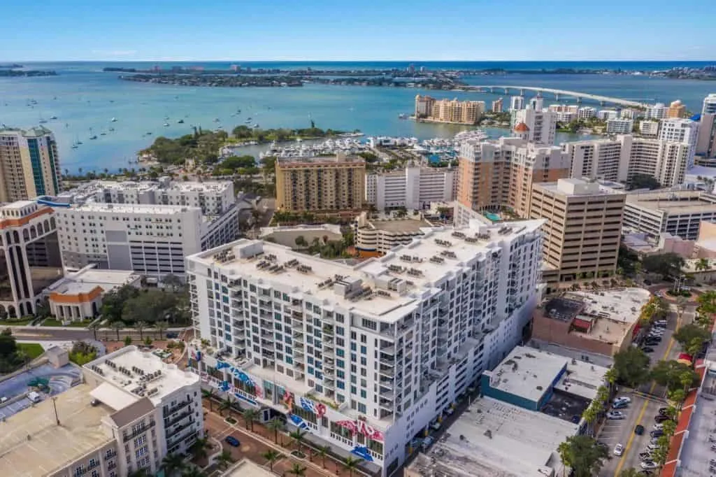 The image showcases an aerial view of a coastal urban area featuring modern buildings and lush greenery, with a serene bay in the background. Boats can be seen on the water, and the skyline includes a mix of residential and commercial structures.
