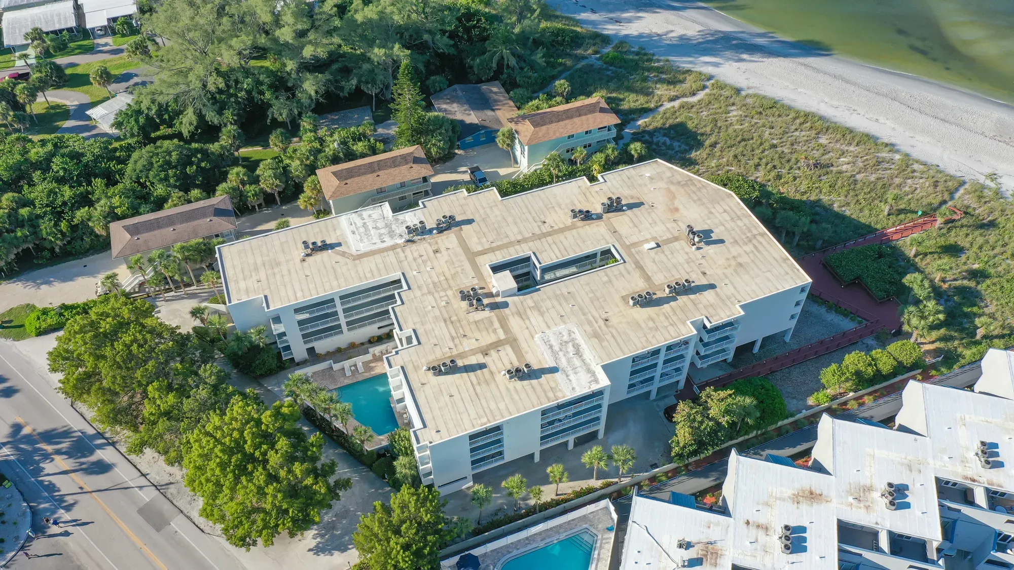 An aerial view of a complex of coastal apartments featuring a flat roof and surrounded by greenery, swimming pools, and a beach nearby. The image captures the layout of the buildings and the proximity to the shoreline.