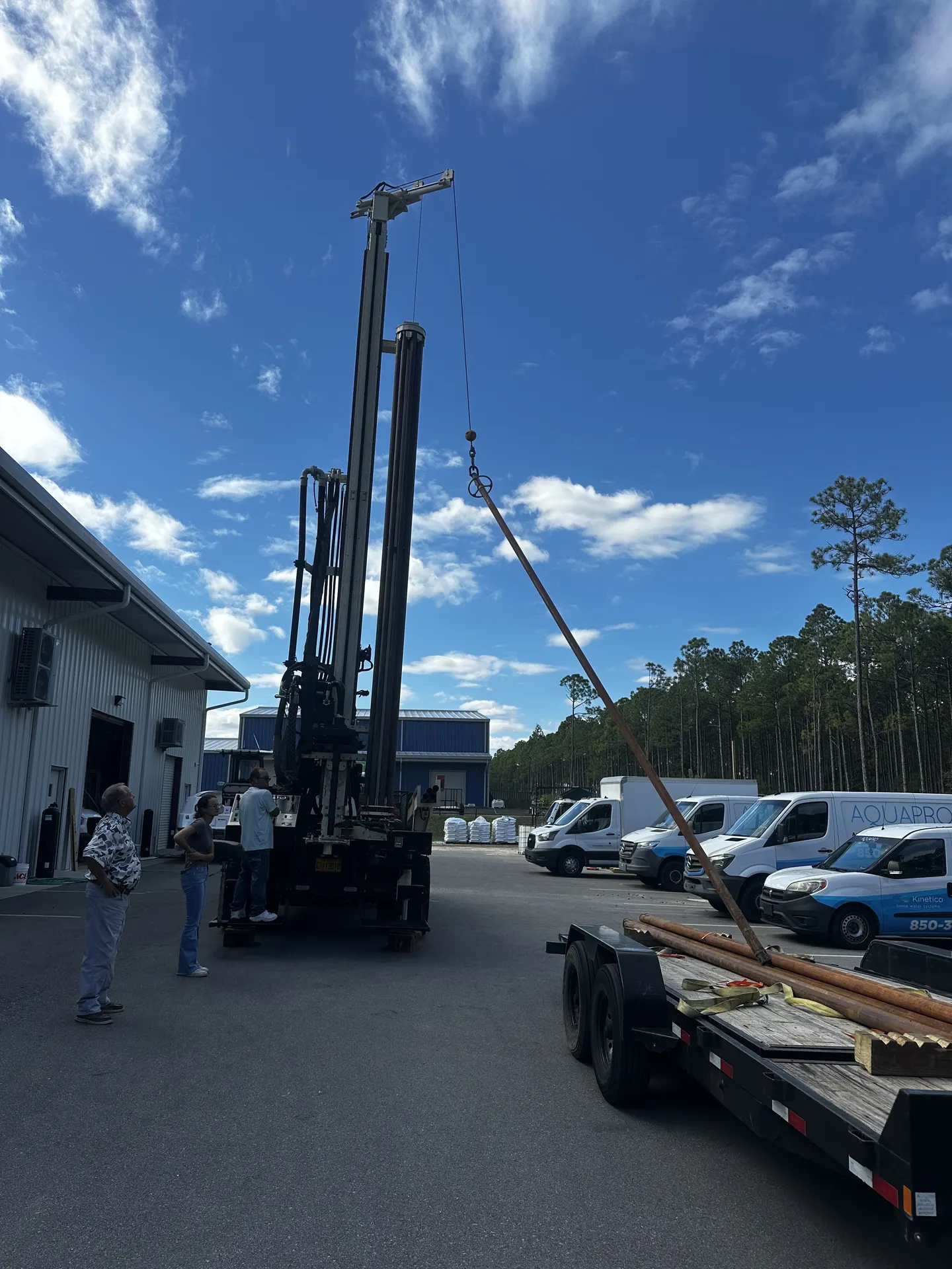 Crew preparing a drilling rig in the yard beside a trailer of casing under a clear blue sky