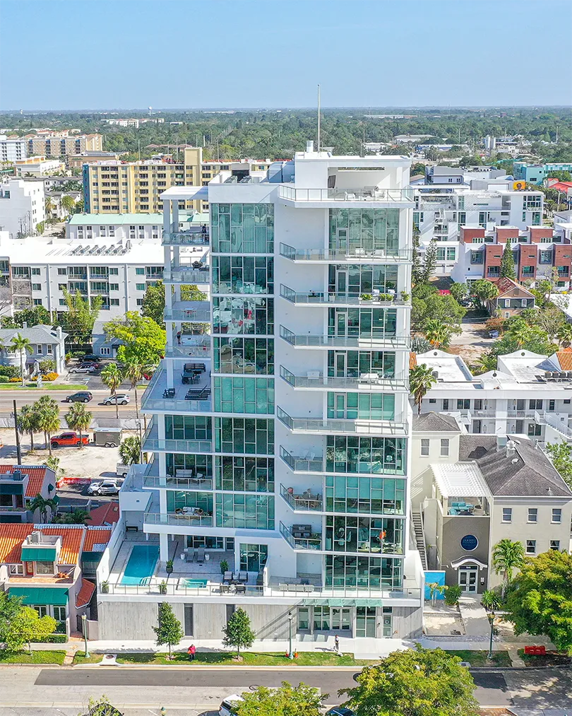 A contemporary glass high-rise building features multiple balconies and a swimming pool on the lower level. Surrounded by residential areas and cityscape, the building showcases modern architecture against a clear blue sky.