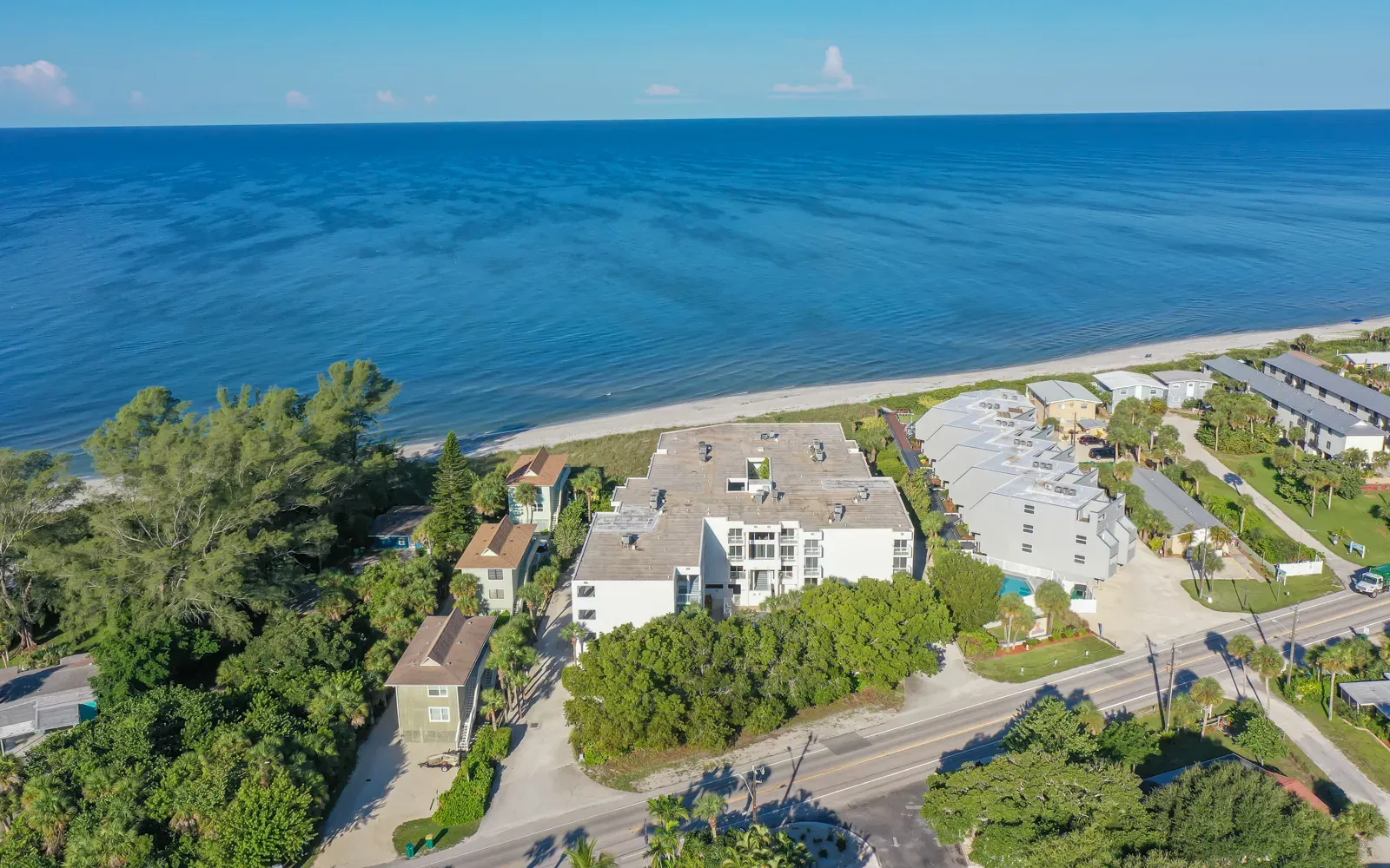 An aerial view showcasing beachfront properties along a serene coastline, with calm blue waters and a sandy beach. Lush greenery and residential buildings complement the picturesque scenery.