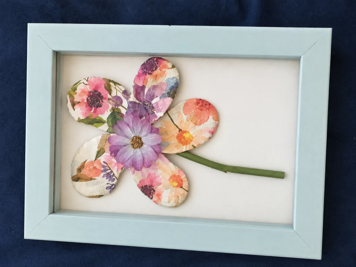 Light blue frame holding a flower made from shells decorated with pink and purple floral prints
