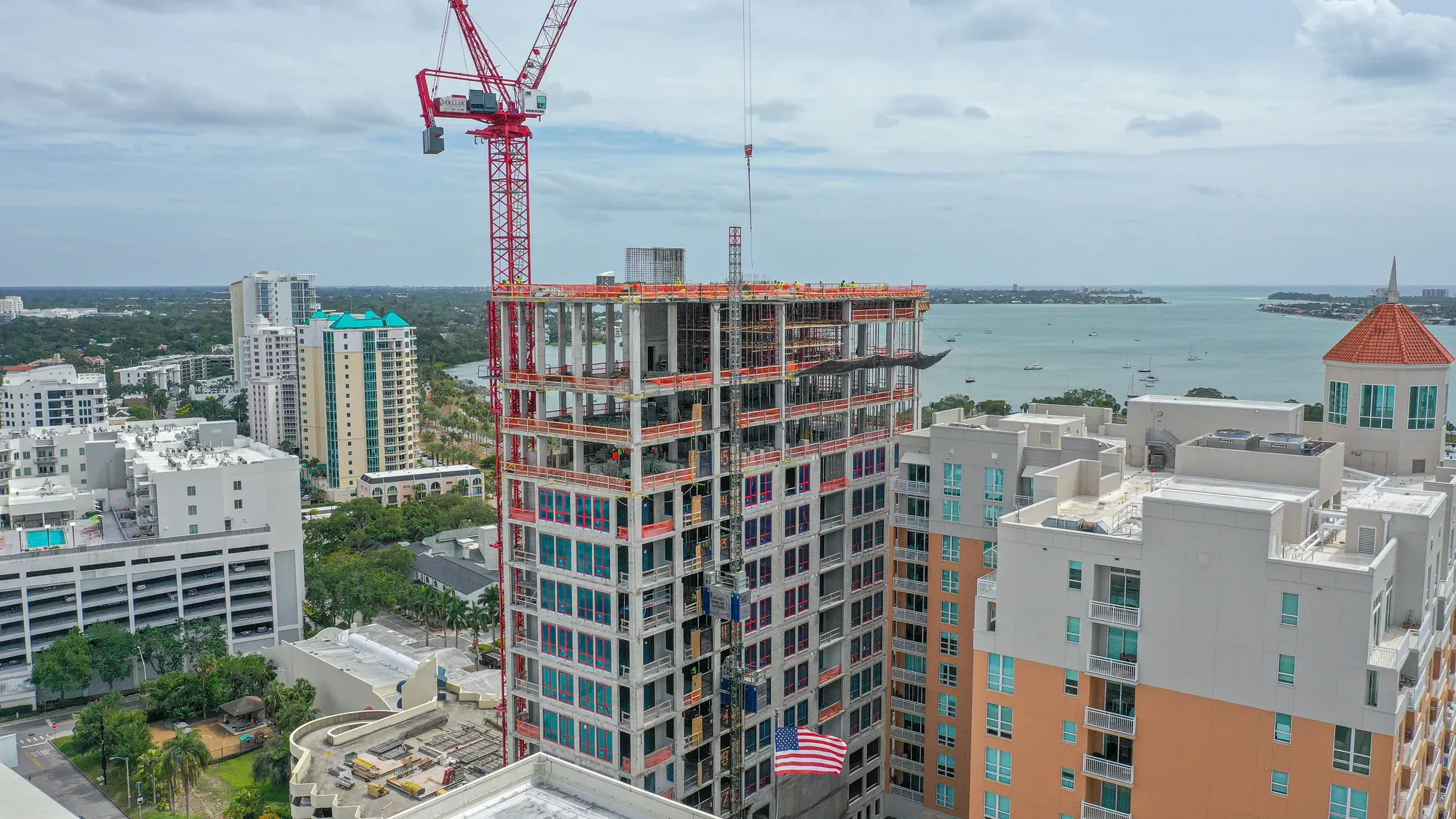 The image showcases an active construction site of a multi-story building, featuring a red crane and sections of concrete structure visible. Surrounding the site are various residential buildings and a scenic view of the waterfront, with boats on the water.