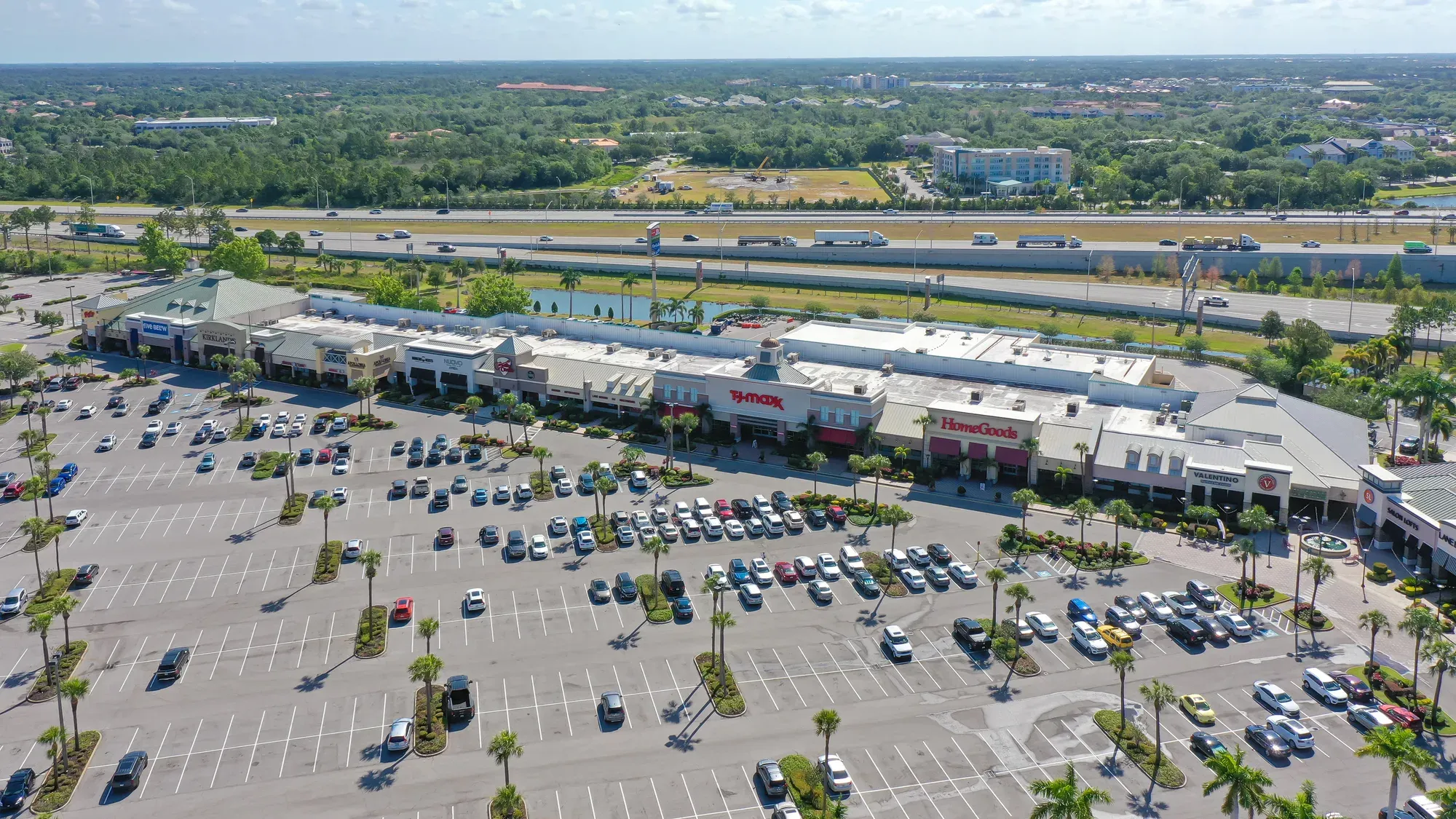 An aerial view of a shopping center featuring various retail stores, including HomeGoods and T.J. Maxx. The expansive parking lot is partially filled with cars, and a highway runs alongside the property.
