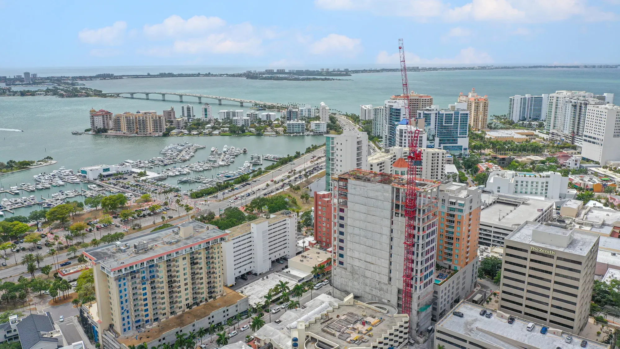 This aerial image captures the Miami skyline featuring a mix of residential and commercial buildings, with a marina and bridge visible in the background. Construction activity is evident with a crane on a building site, showcasing the city's ongoing development.