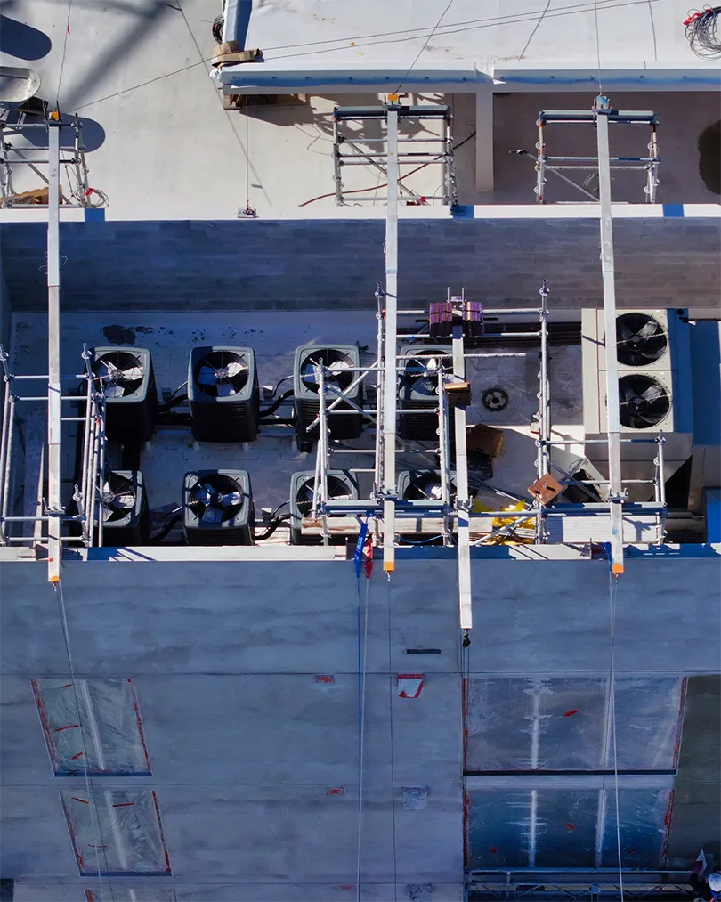 An aerial view of a construction site showing an array of rooftop air conditioning units and scaffolding. The image captures unfinished concrete walls and construction materials, indicating ongoing building work.